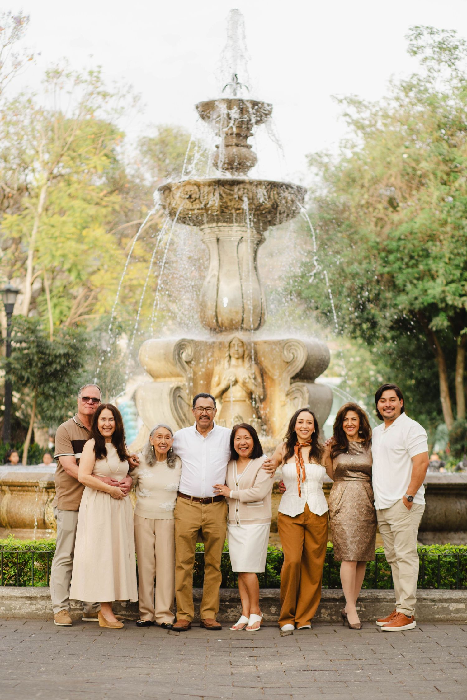 A group poses together in casual beige and brown outfits by a stone fountain in an outdoor garden setting.