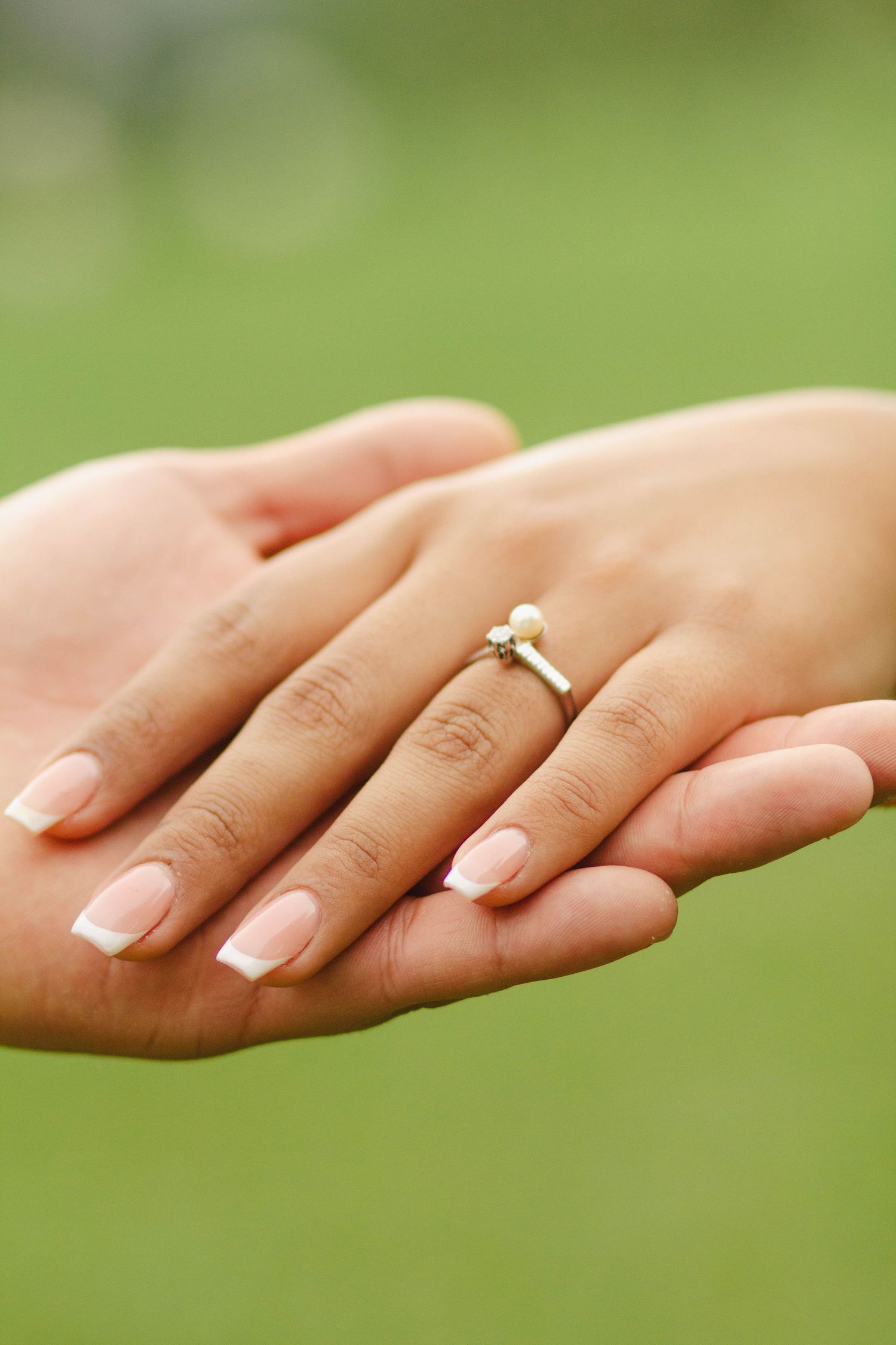 Close-up of manicured hand displaying delicate engagement ring against blurred green background.