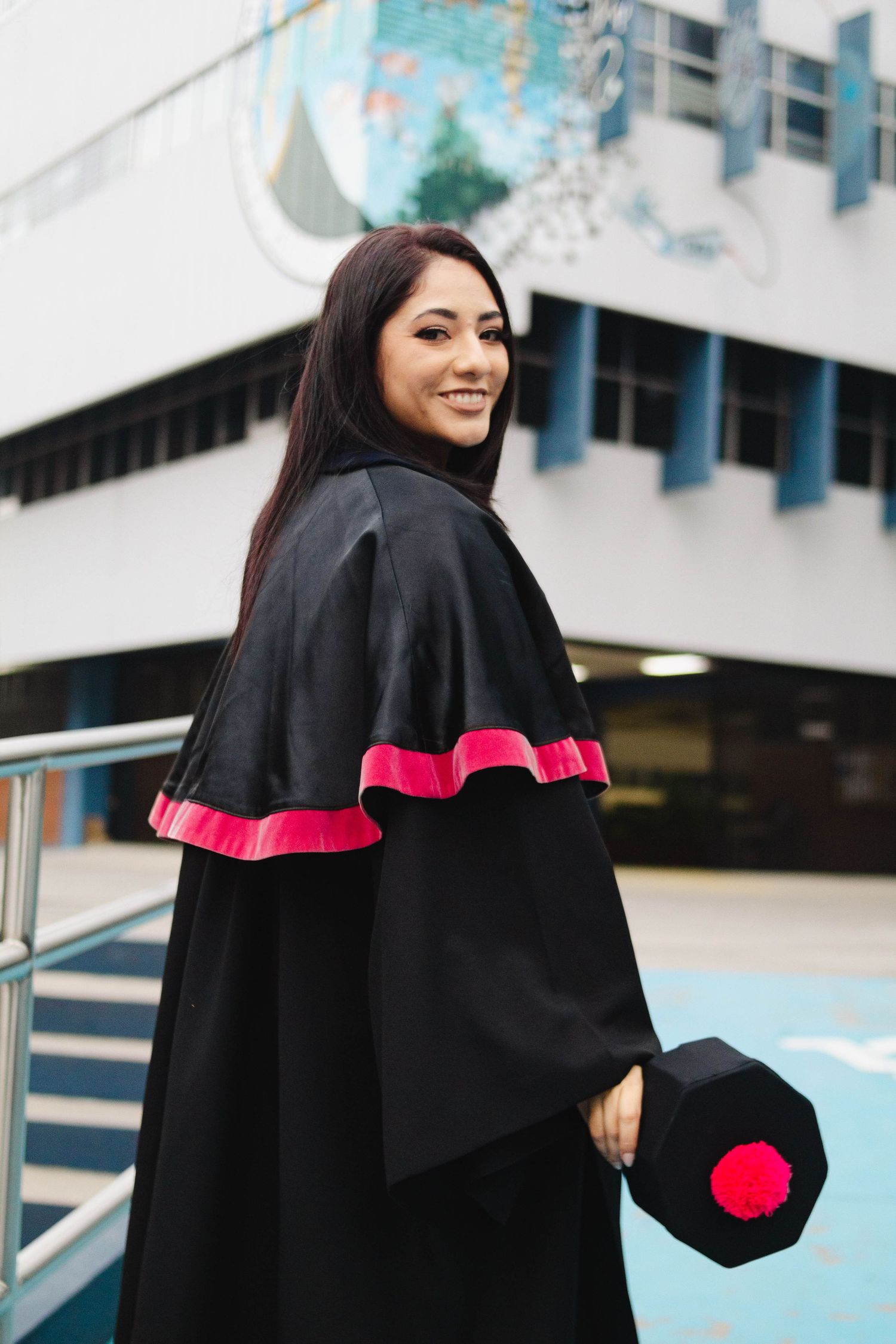 Graduate in black and pink academic regalia poses outside modern building with graduation cap.