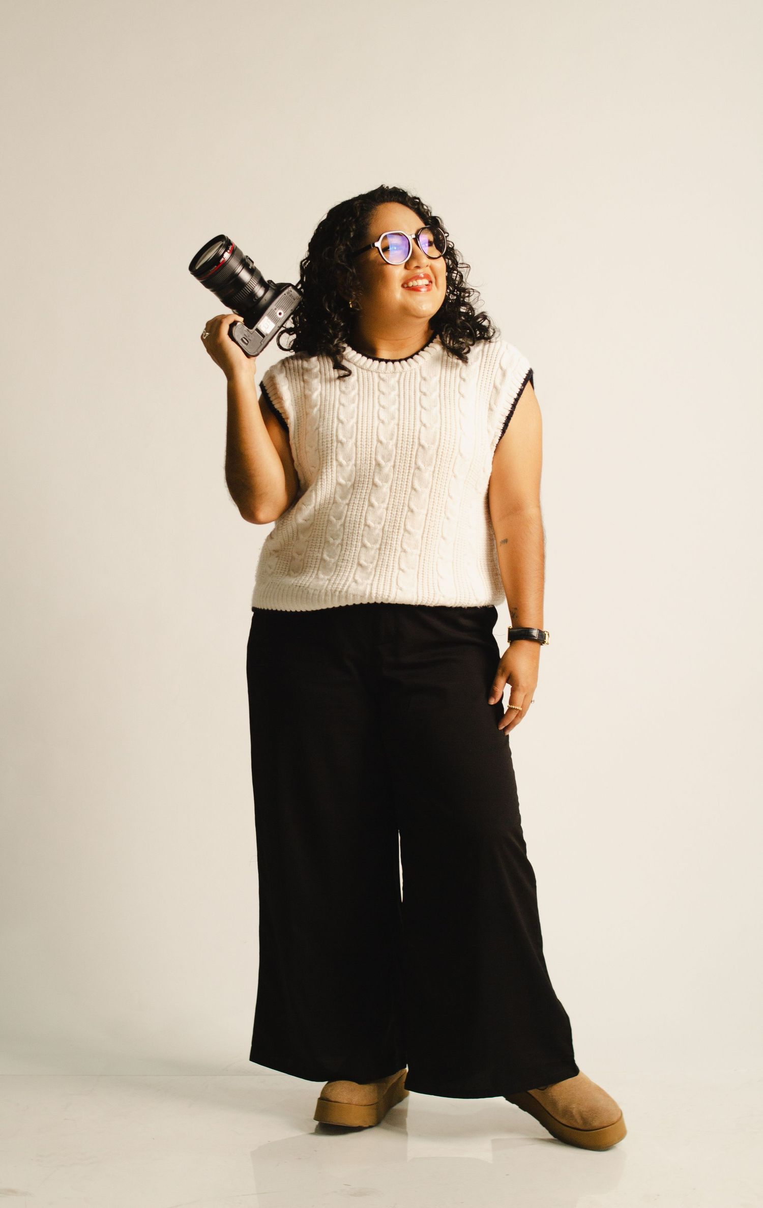 Person in cream knit vest and black palazzo pants holds camera while posing against beige background.