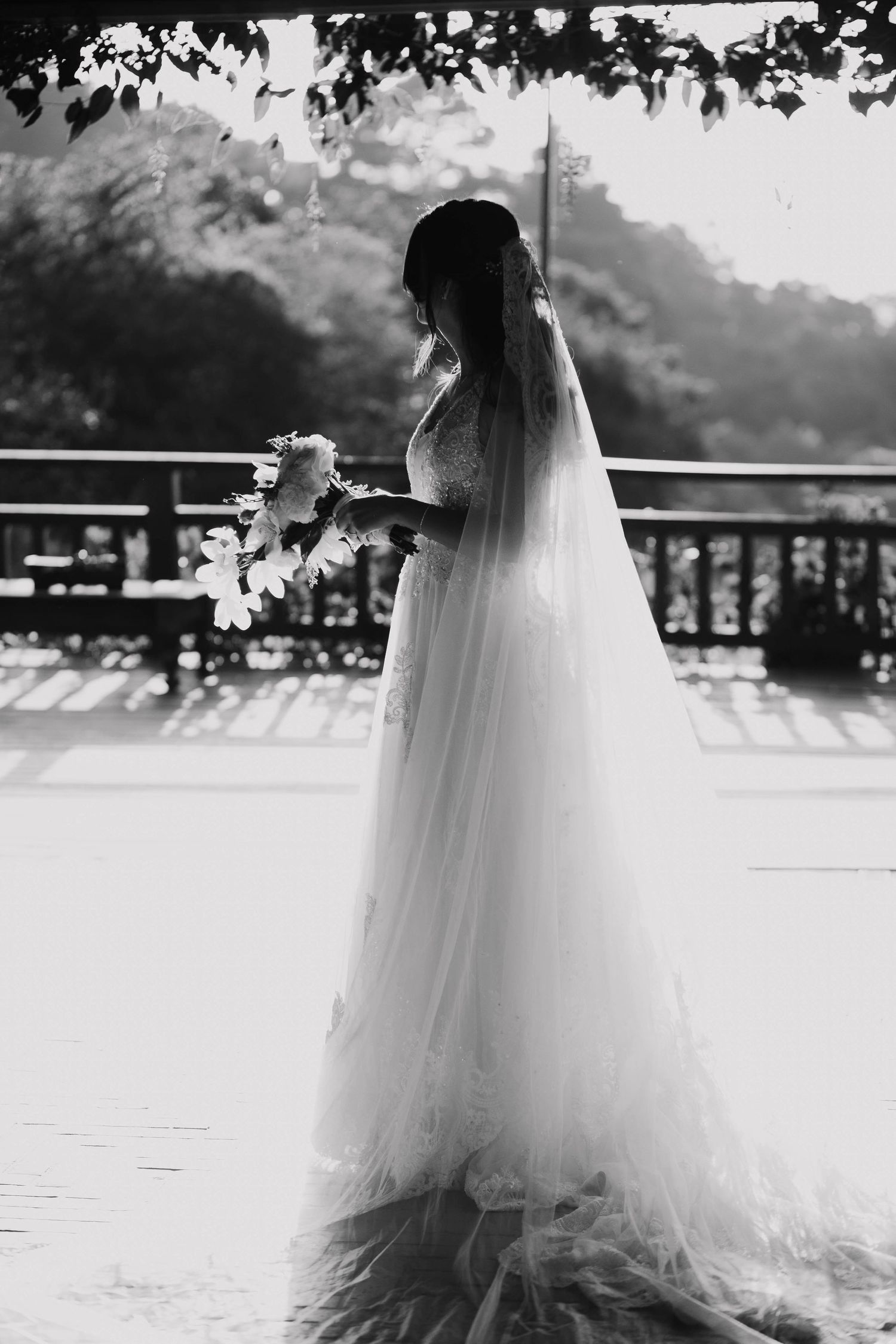 Silhouette of a bride in flowing veil standing on a balcony holding a bouquet.