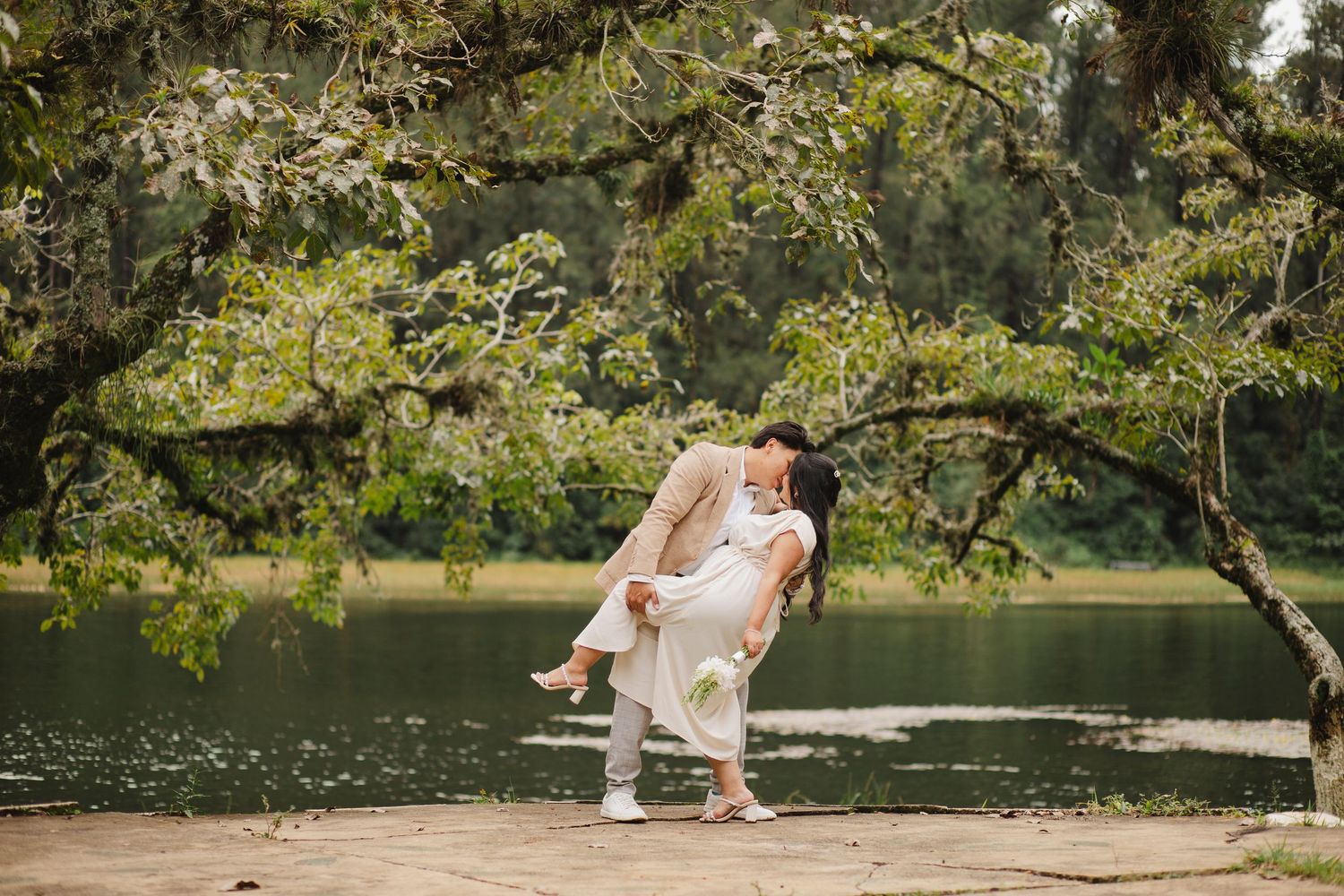 A romantic couple lounges on a blanket by a serene lake surrounded by lush greenery on a sunny afternoon.