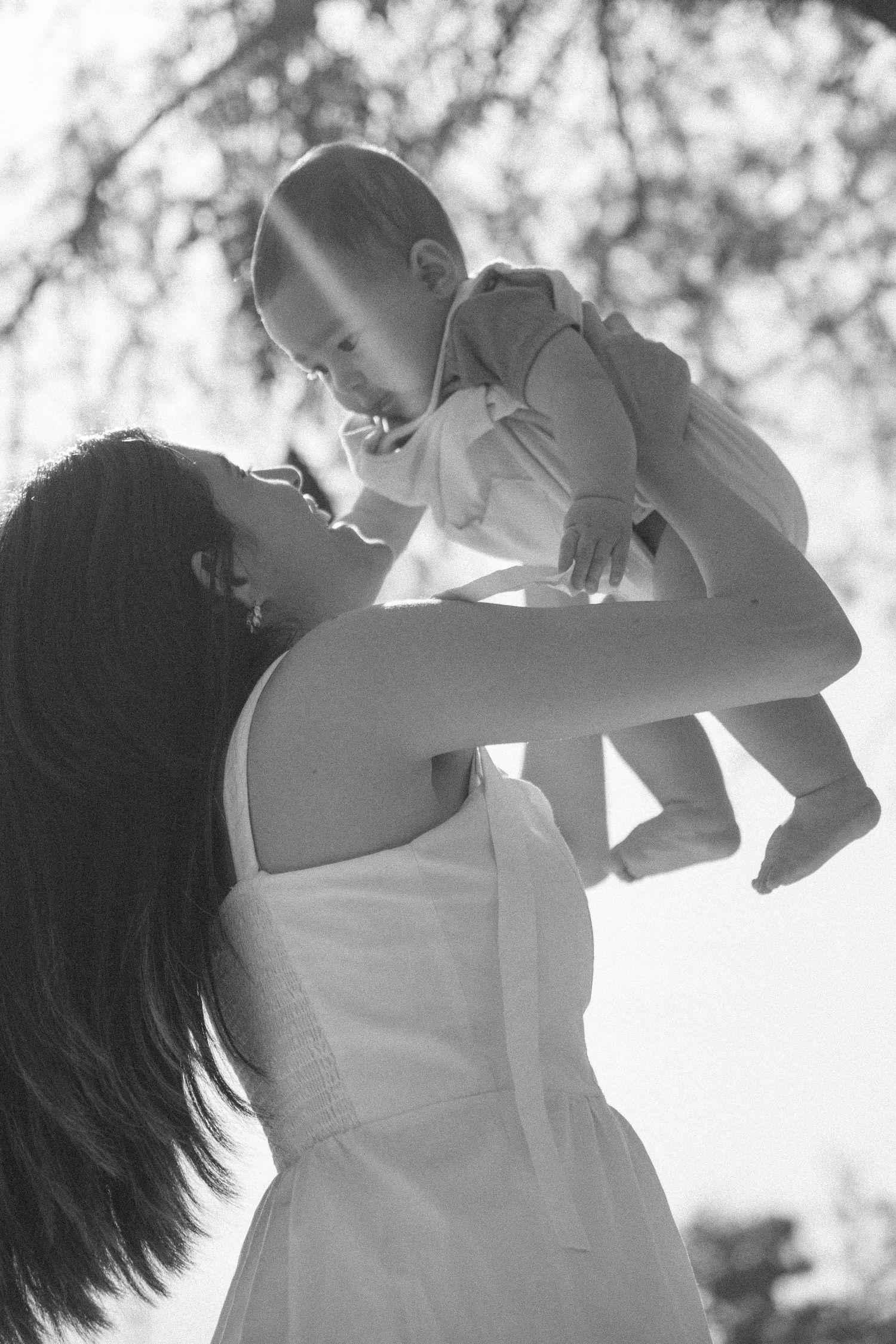 Black and white photo of a mother lifting her baby in the air outdoors, sharing a tender moment.