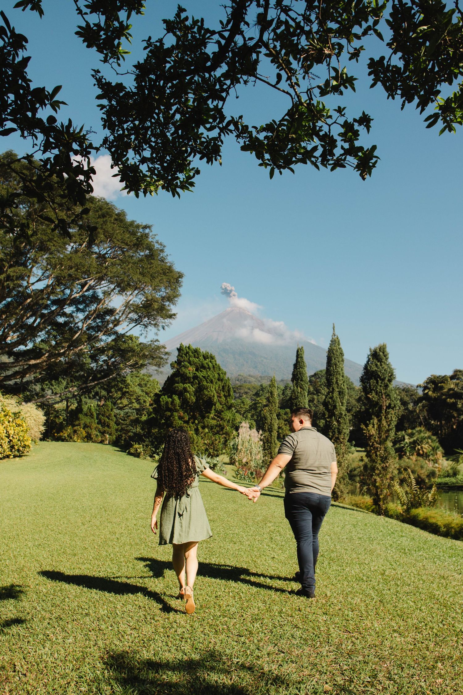 A couple walks hand in hand across a green lawn with a volcanic mountain visible in the distance.