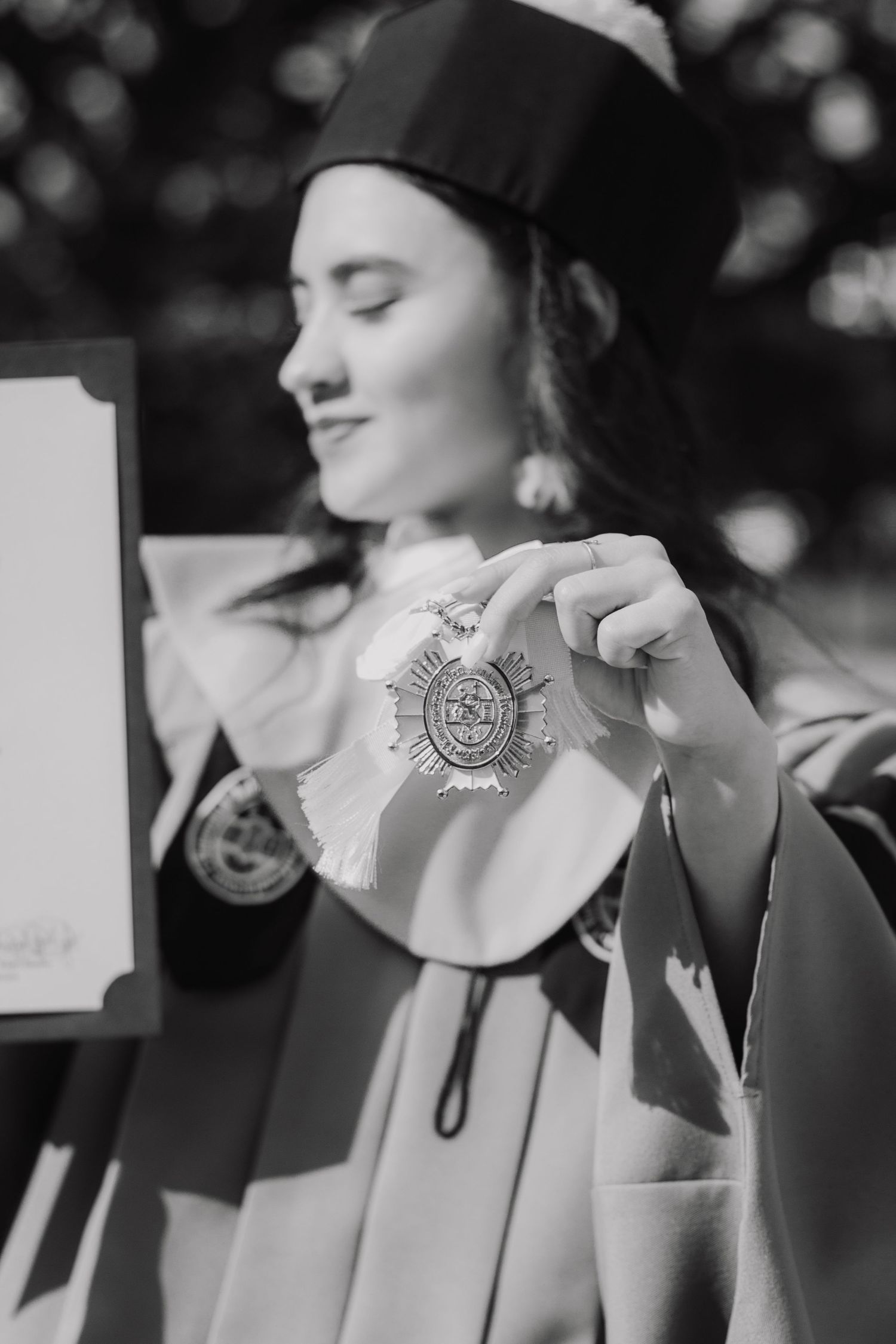 Graduate holding academic medallion while wearing graduation cap in monochrome photo.
