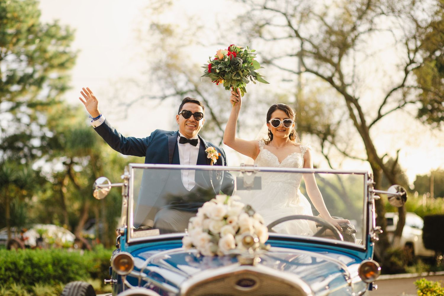 Happy couple celebrating in vintage blue convertible car with bouquet raised in air.
