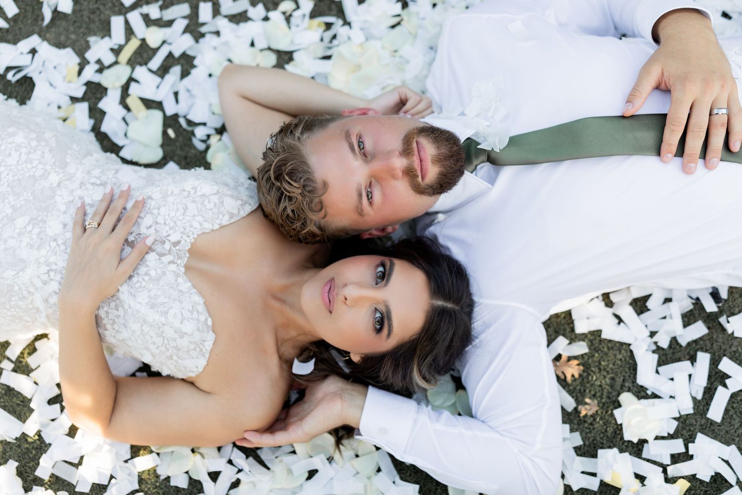 A couple in white wedding attire lies together on the ground surrounded by scattered autumn leaves.