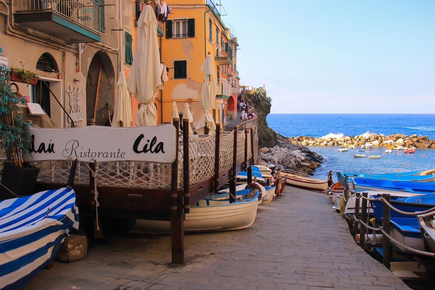 Traditional boats and blue-striped umbrellas line a scenic coastal walkway in an Italian village.