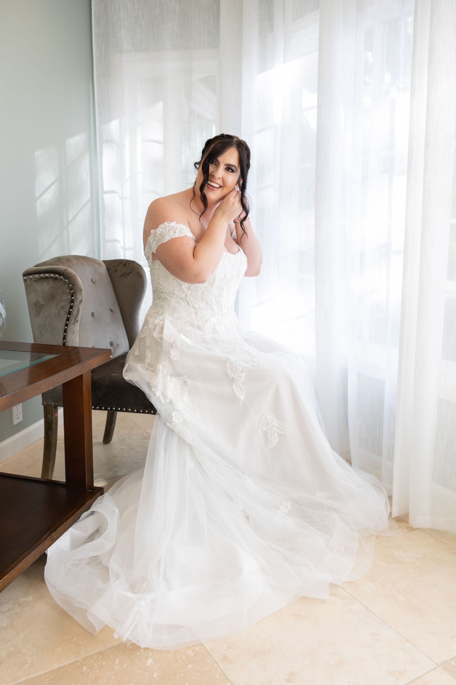 A stunning off-shoulder white wedding gown with delicate lace details and flowing train photographed in a bright interior setting.