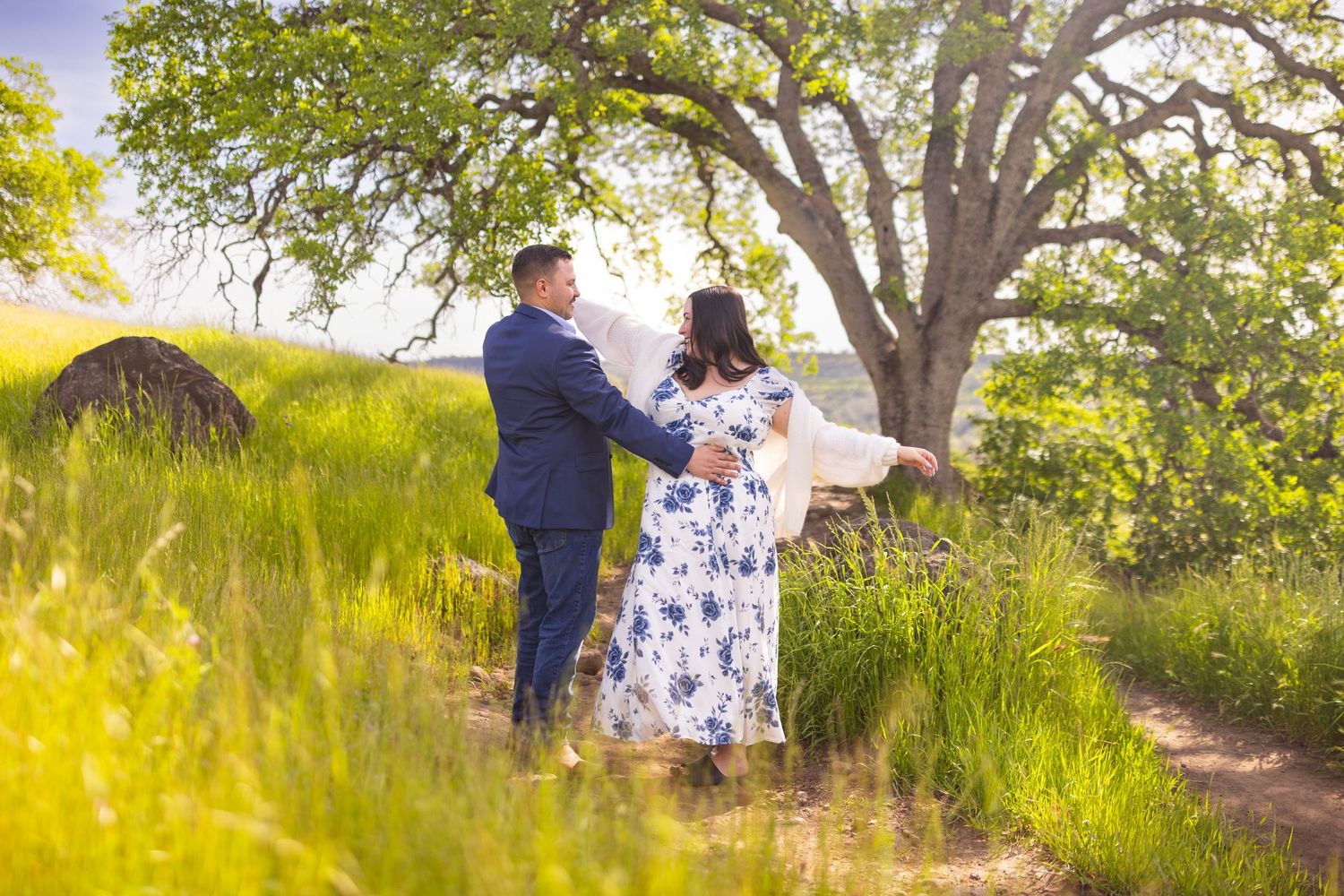 A couple embraces in a romantic moment under a large tree during golden hour in a sunlit meadow.