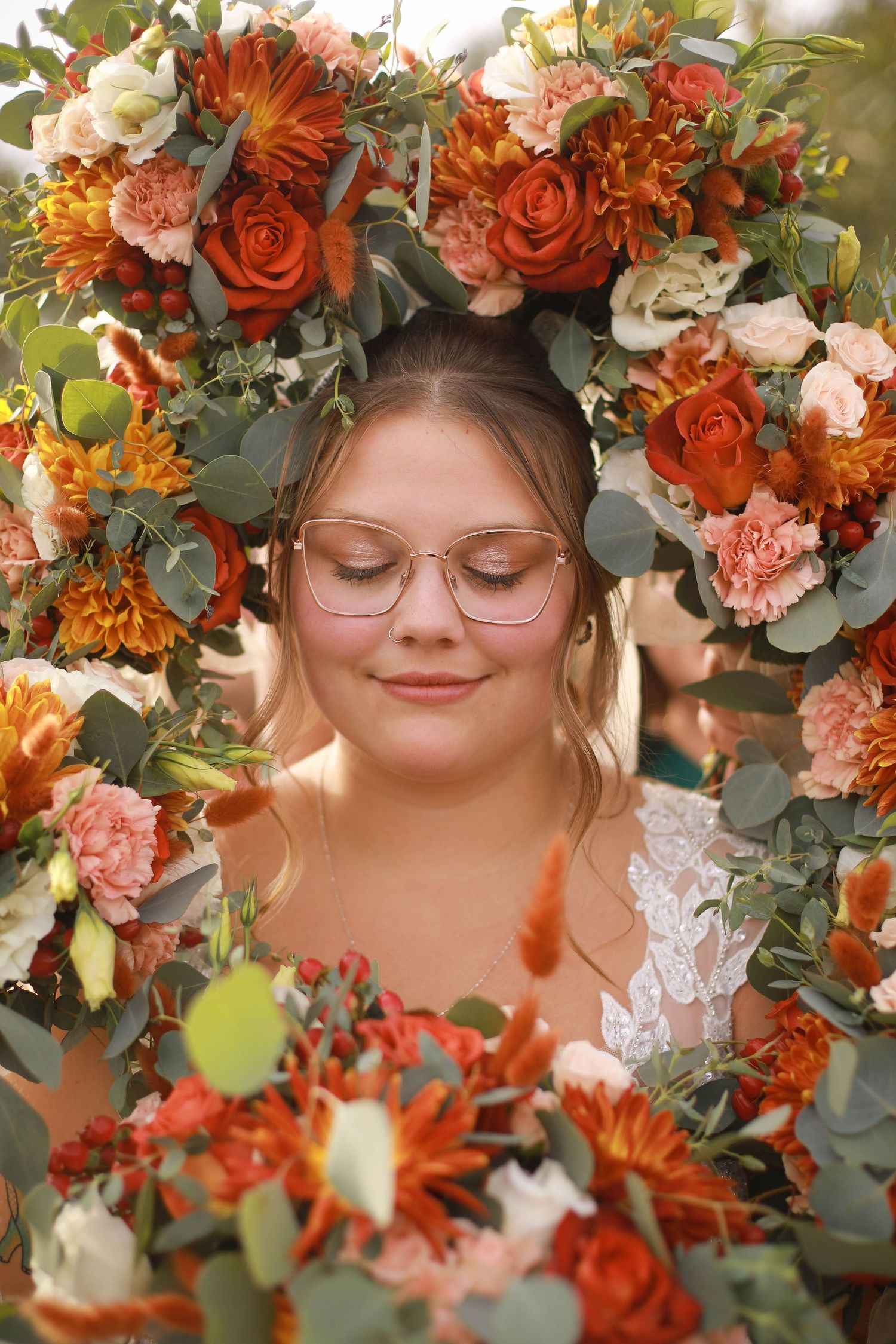 A person wearing glasses smiles peacefully surrounded by a crown of orange and pink flowers.