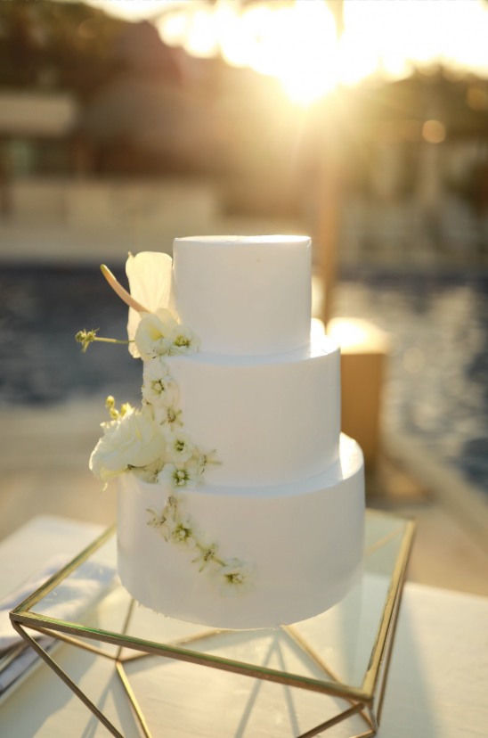 Elegant three-tier white wedding cake decorated with orchids sits on mirrored stand during golden sunset.