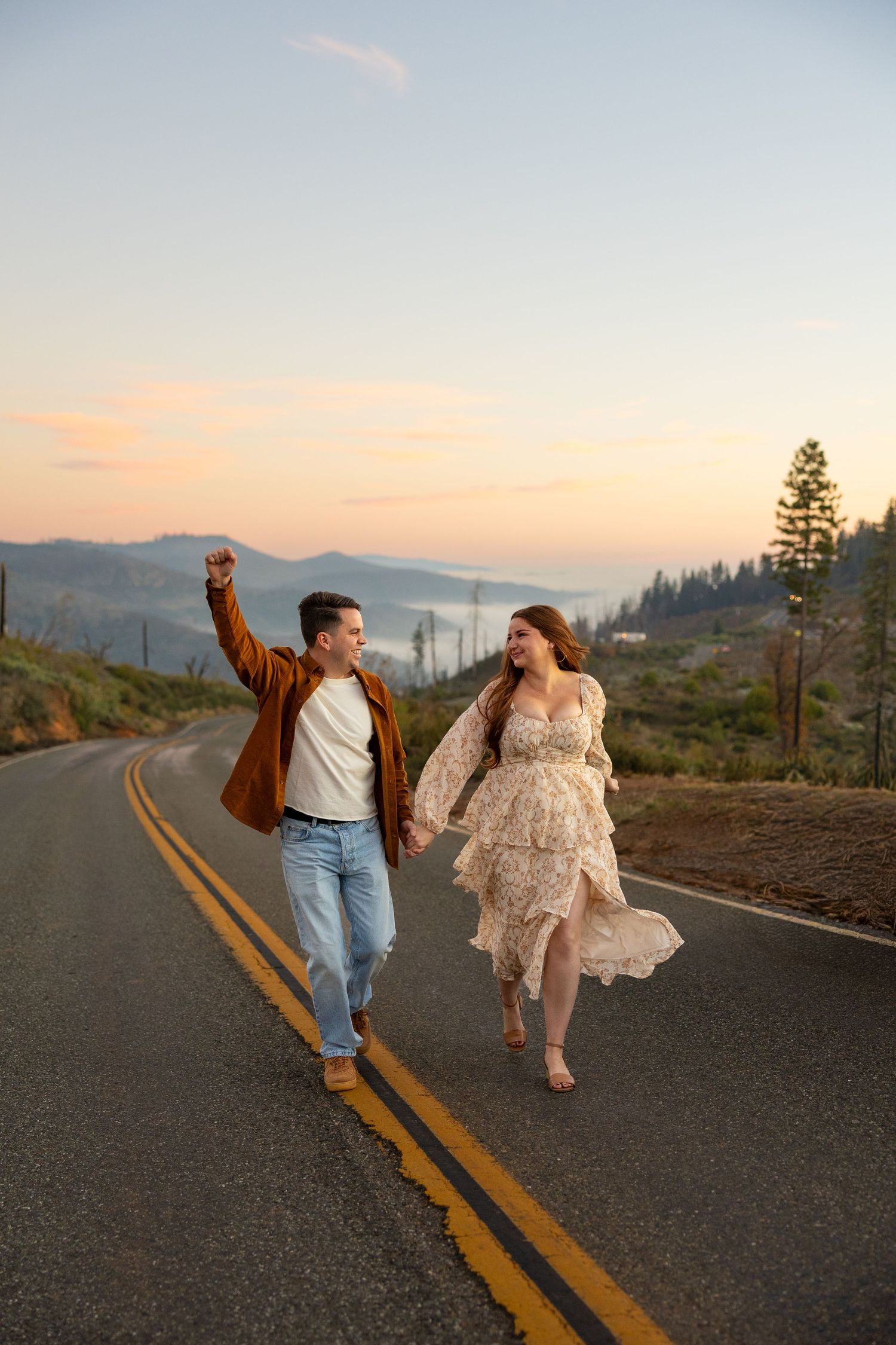 Couple dances playfully on a mountain road at sunset with scenic views.