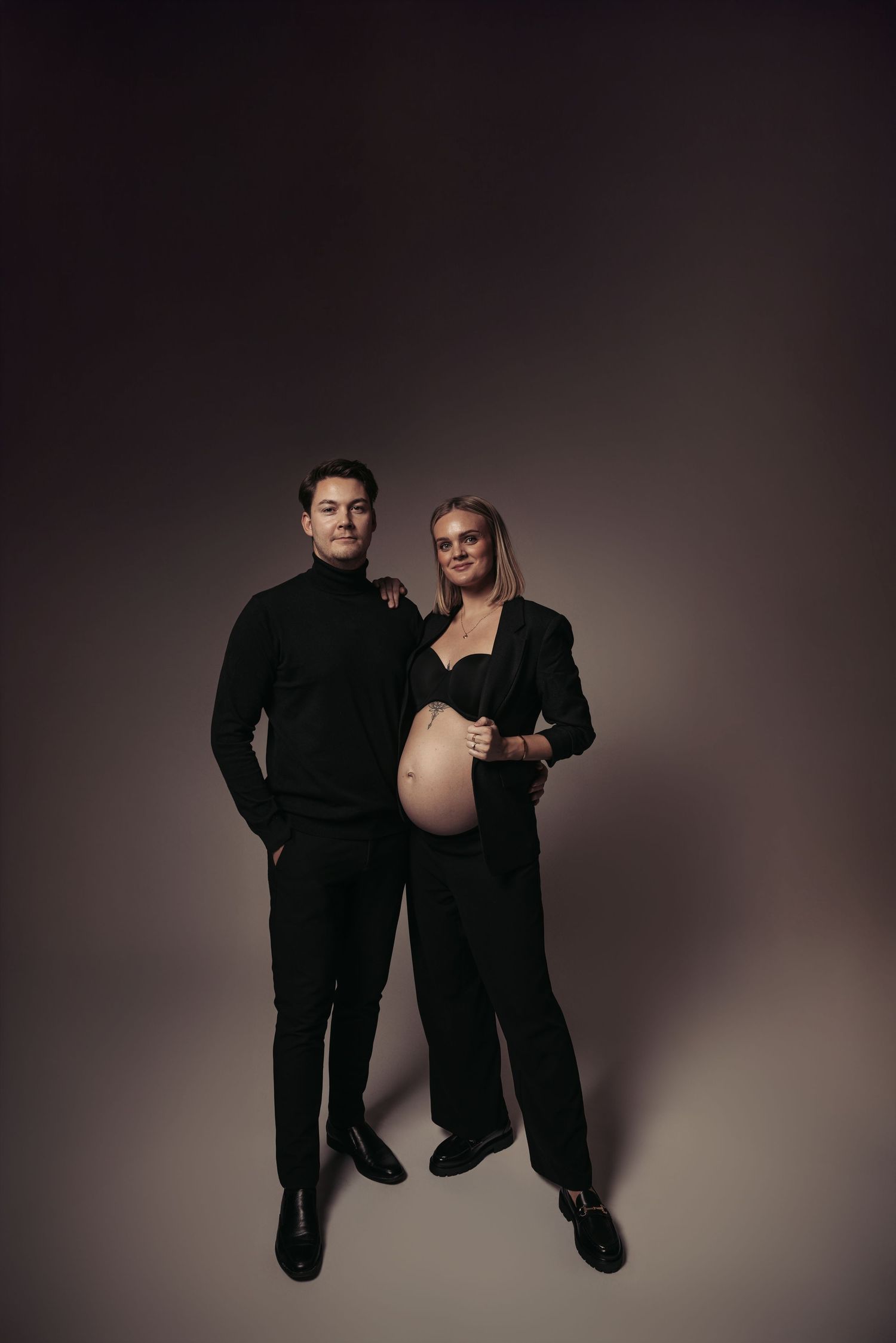 Couple in black outfits posing for maternity photo, pregnant woman showing baby bump, studio dark background.