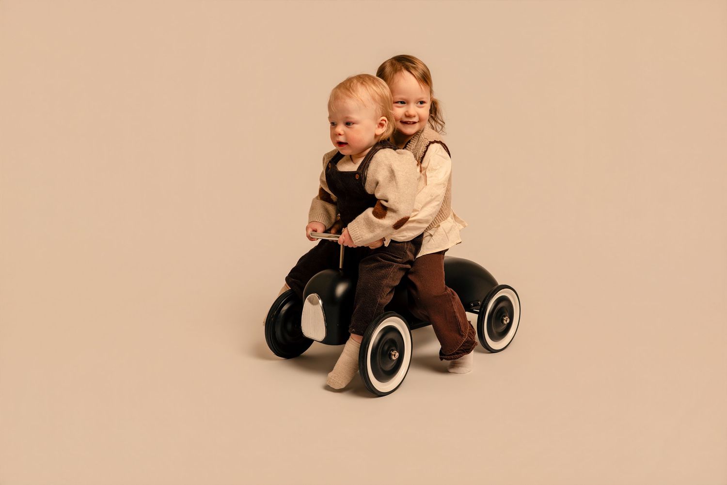 Two toddlers share a ride on a black balance bike toy against a warm beige background.