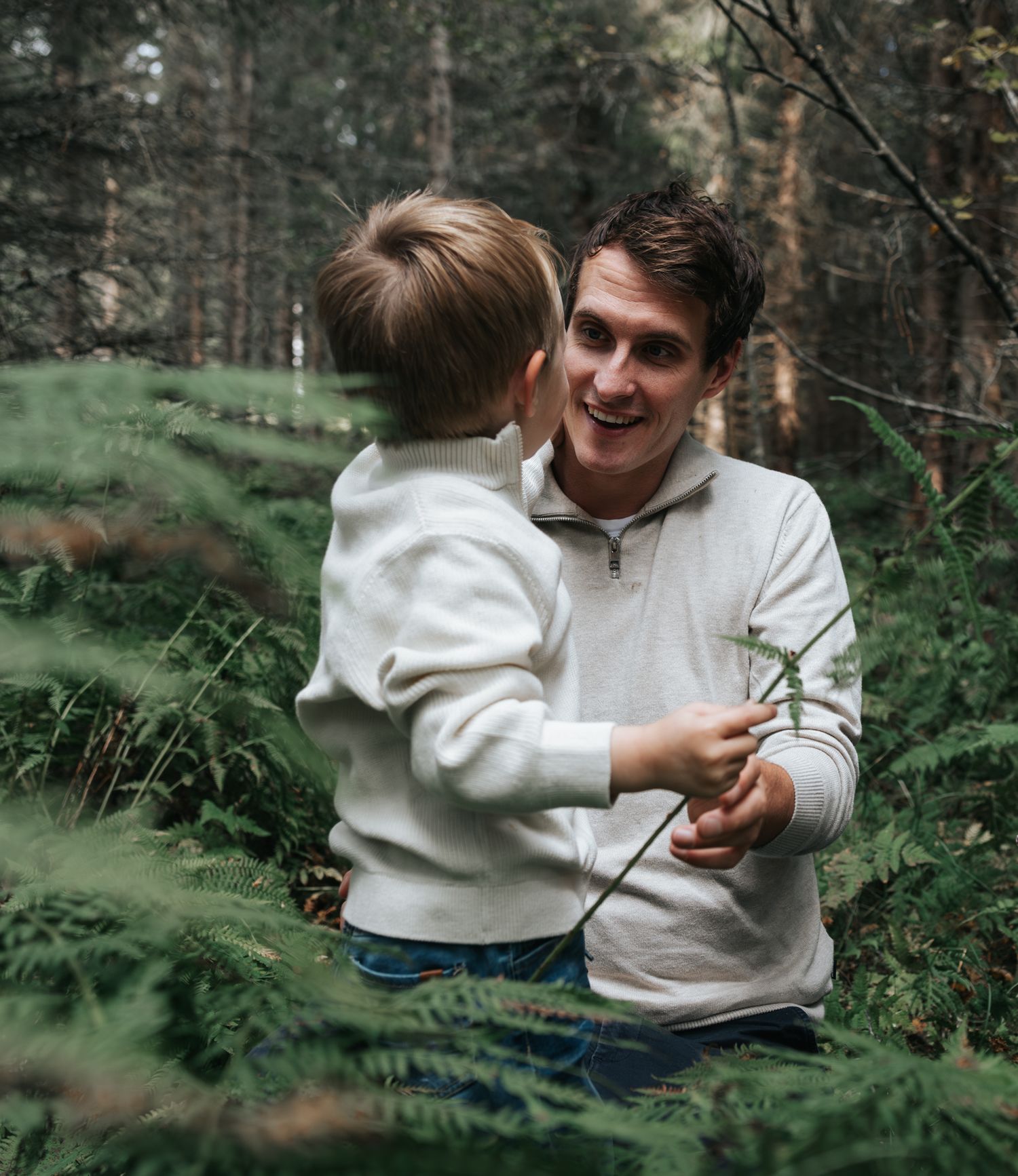 Smiling father kneeling to interact with young son among green ferns in a forest.