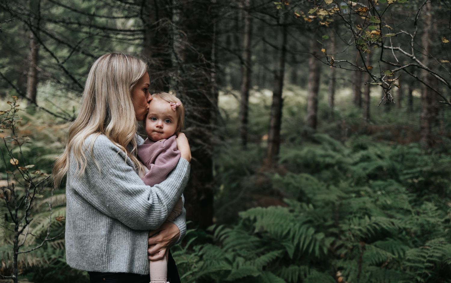Blonde mother tenderly holding young toddler girl in pink sweater in a forest setting.