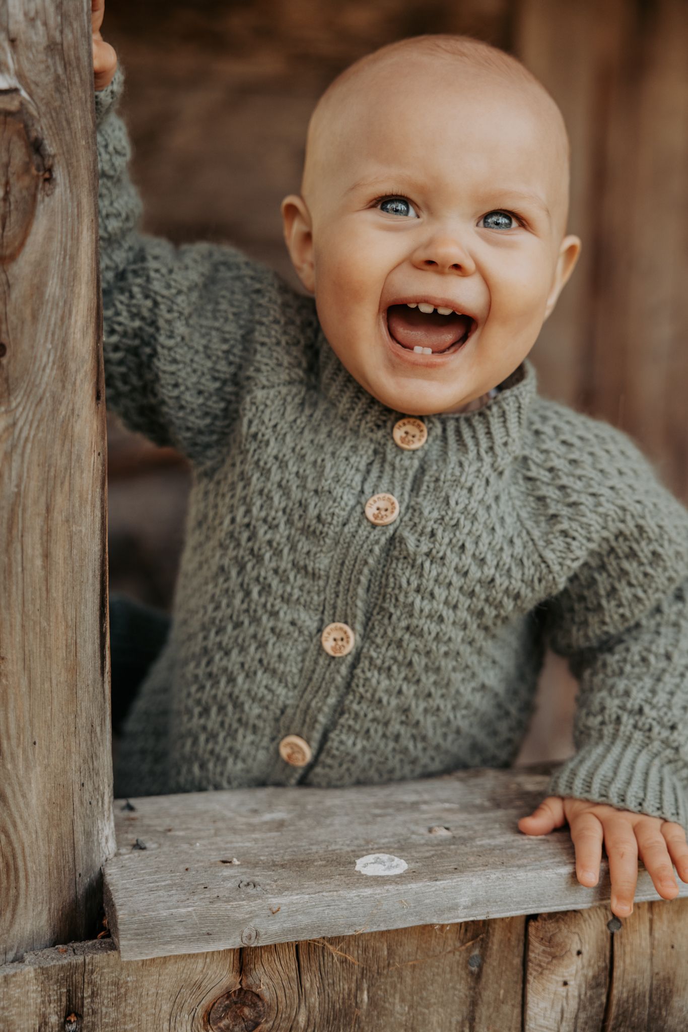 Happy laughing baby in green knit cardigan stands at a rustic wooden fence outdoors.
