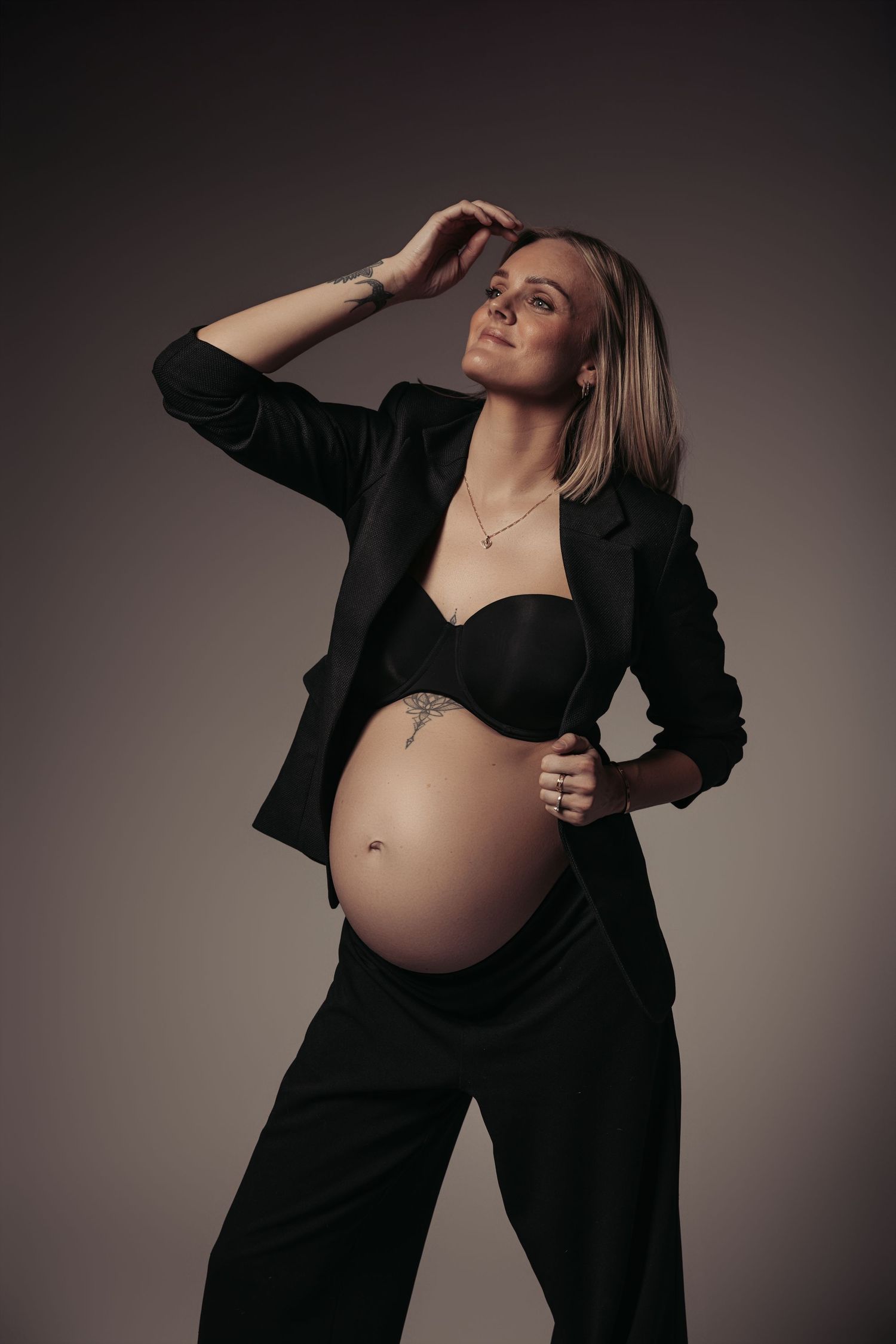 Pregnant woman in black blazer and bra posing confidently in studio, showcasing baby bump.