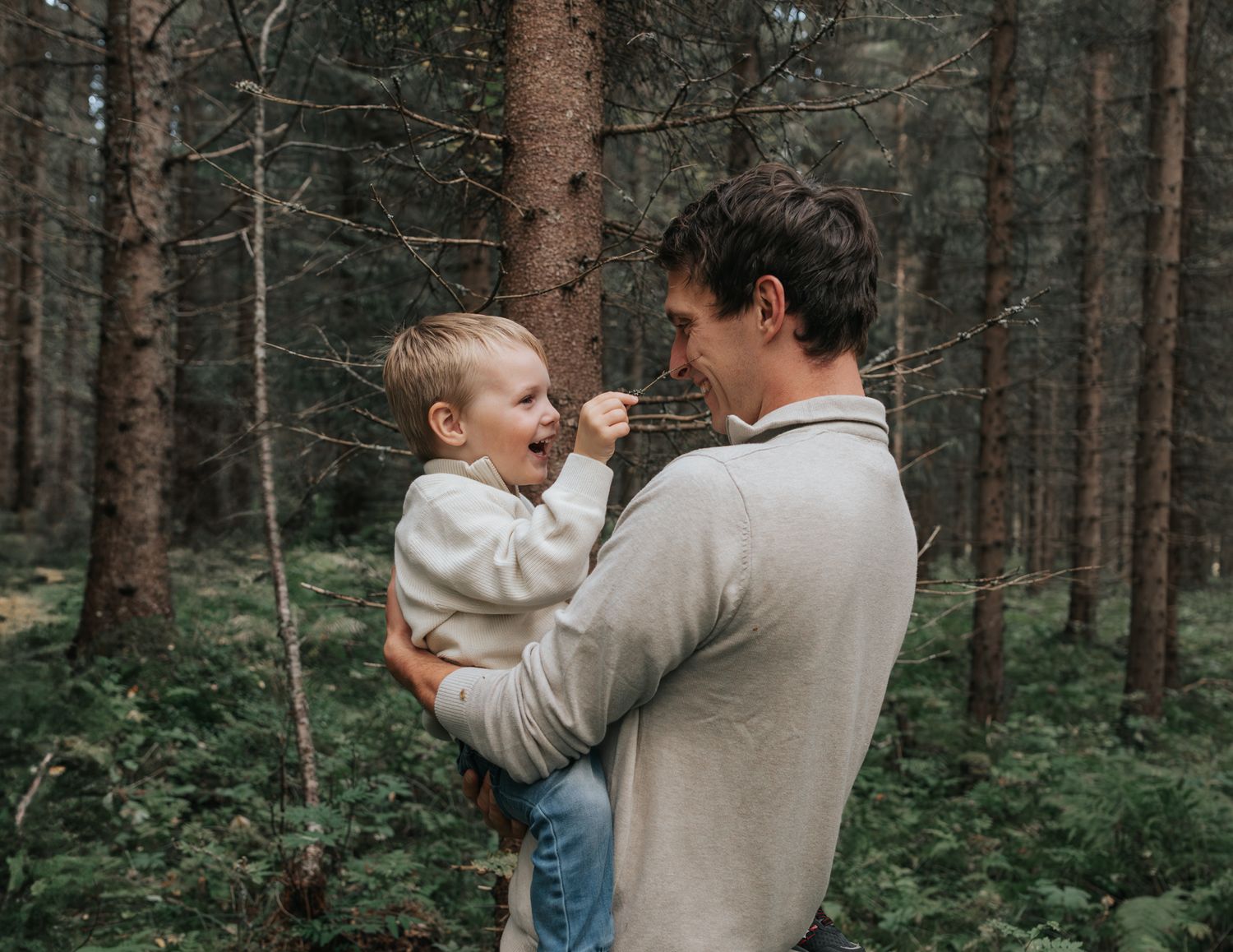 Father holding laughing toddler boy in a lush green forest setting.