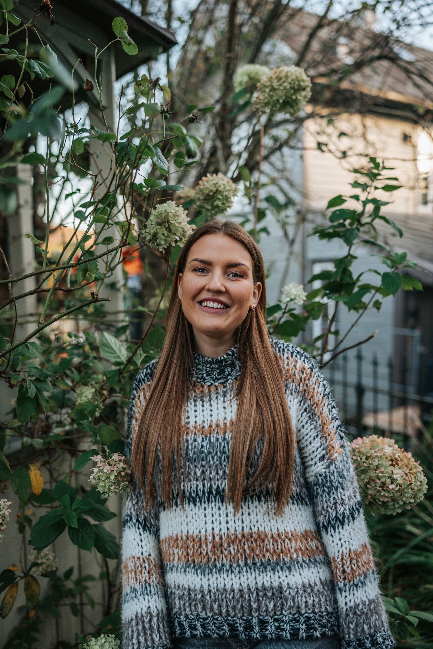 Woman with long brown hair wearing a striped knit sweater smiling in a garden with greenery and flowers.