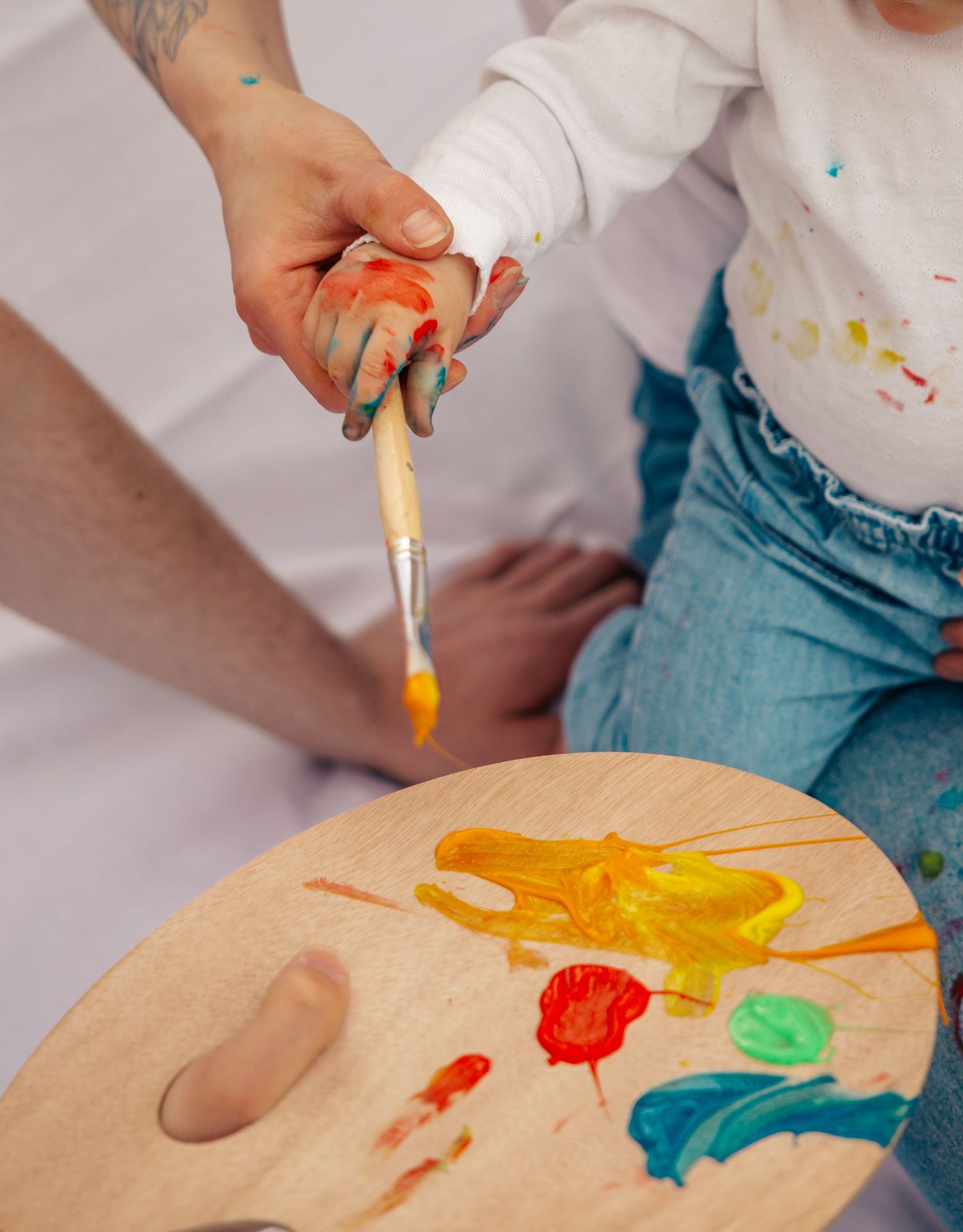 An adult guiding a toddlers hand with a paintbrush over a colorful wooden palette.