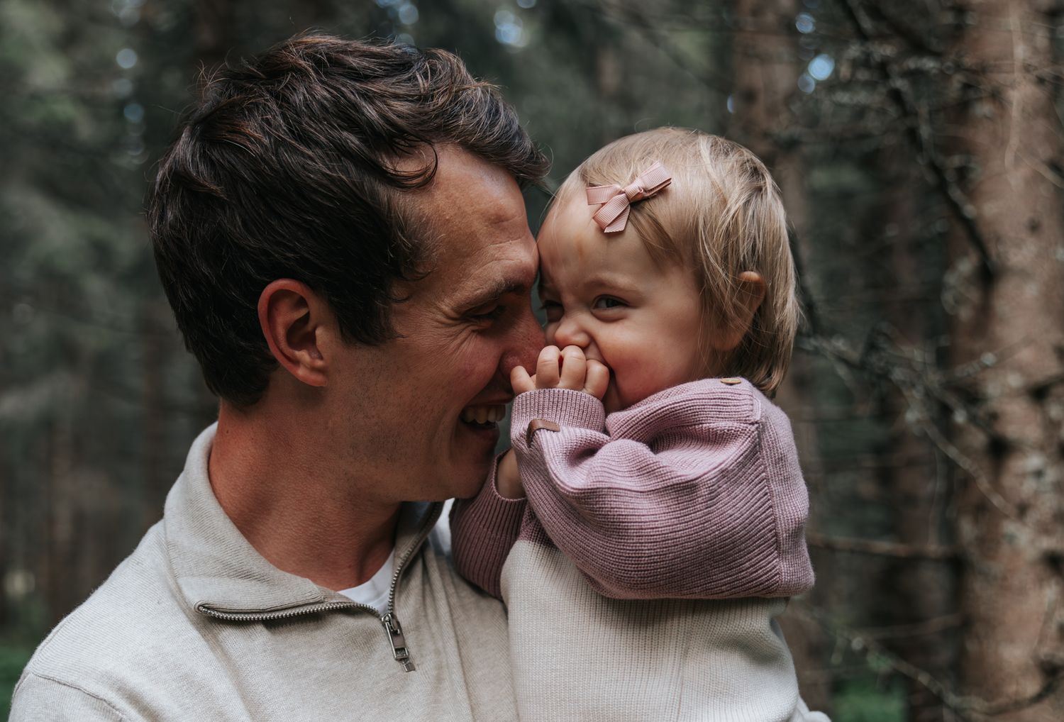 Father cheek-to-cheek with smiling baby girl in a forest backdrop.