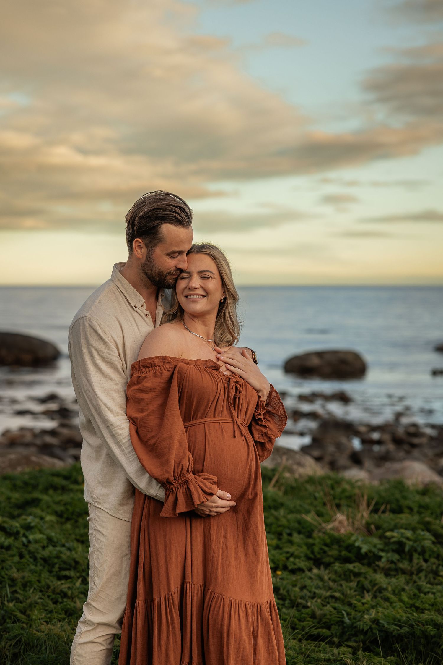 Couple embracing at sunset by rocky ocean shore, pregnant woman in rust boho dress smiling joyfully during maternity photoshoot.