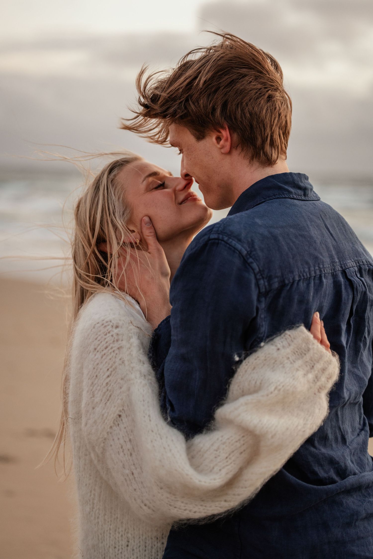 Young couple about to kiss while embracing on a windy beach at dusk.