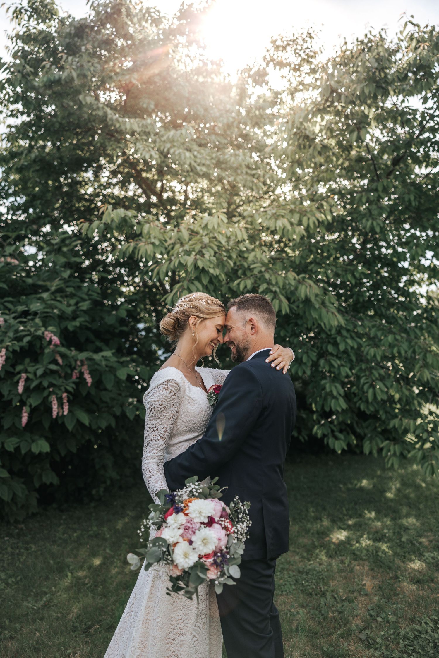 Bride and groom touching foreheads tenderly in sunlit garden, bride in lace dress holding vibrant bouquet.