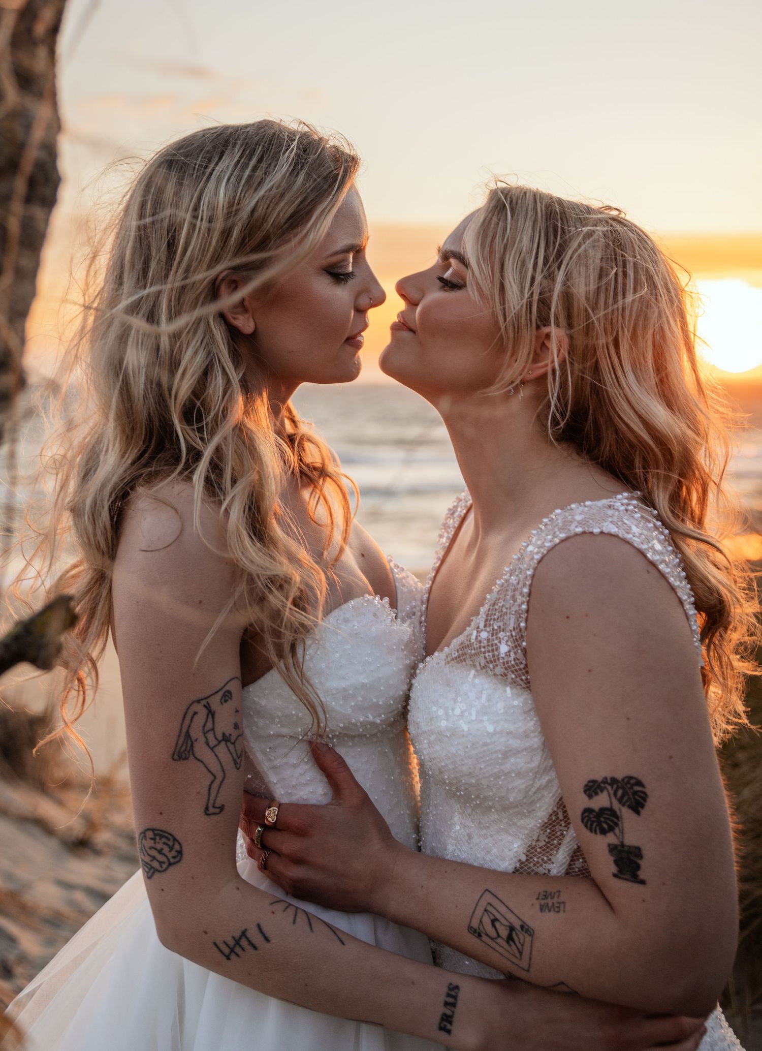 Two brides in white gowns sharing a kiss at sunset beach, both with blonde hair and tattoos.