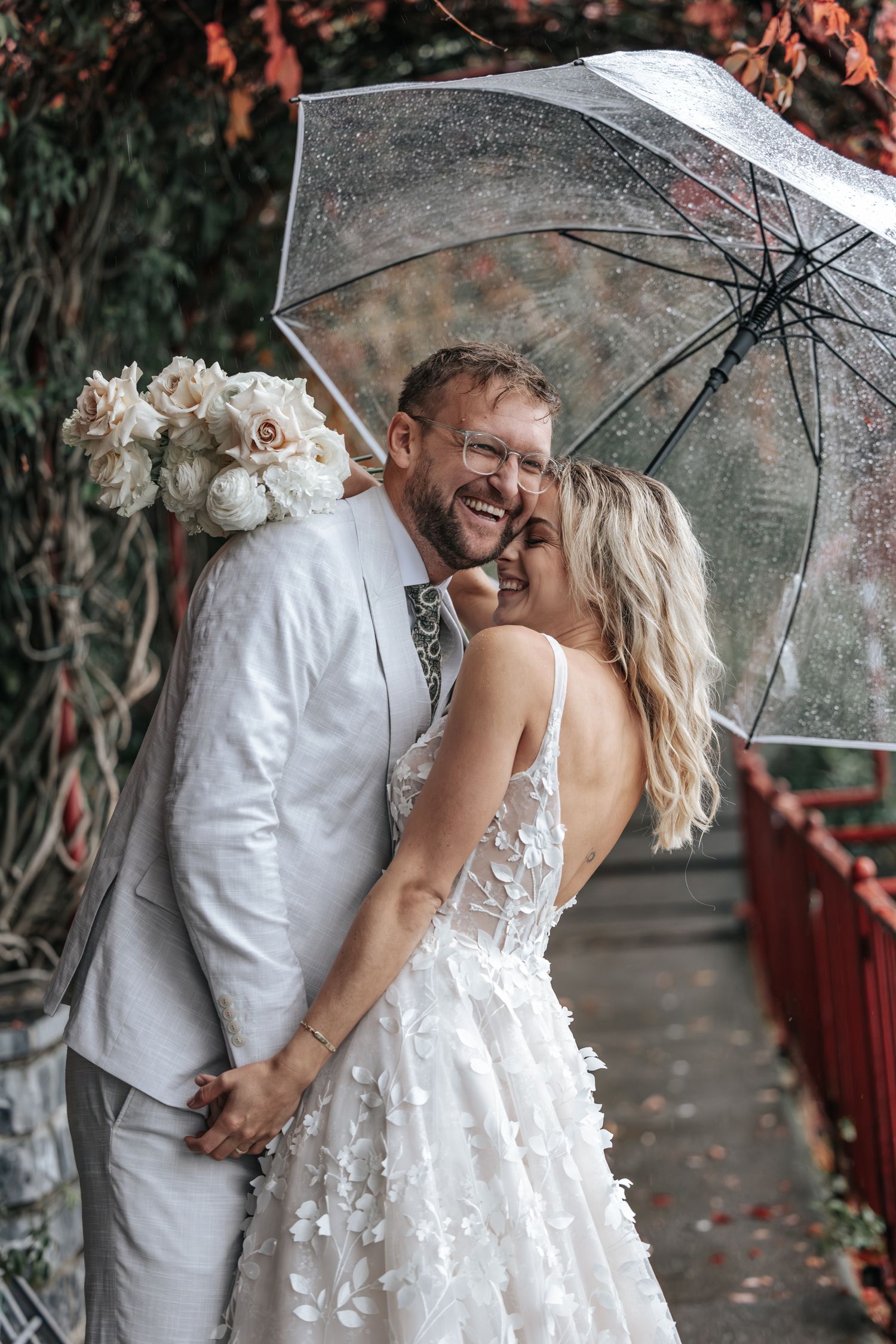 Joyful bride and groom laughing under a clear umbrella surrounded by red autumn foliage on their wedding day.