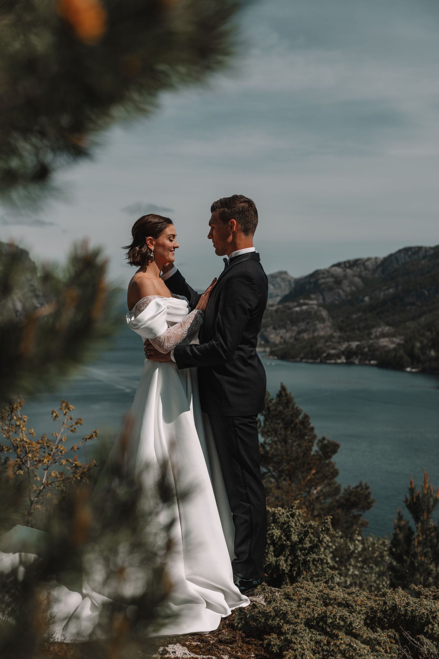 Elegant bride and groom embrace on a rocky overlook with stunning fjord and mountain views behind them.