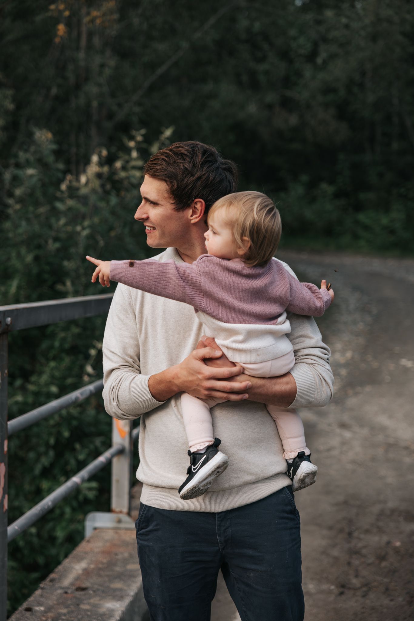 Father holding baby girl in pink sweater while pointing outdoors near a bridge.