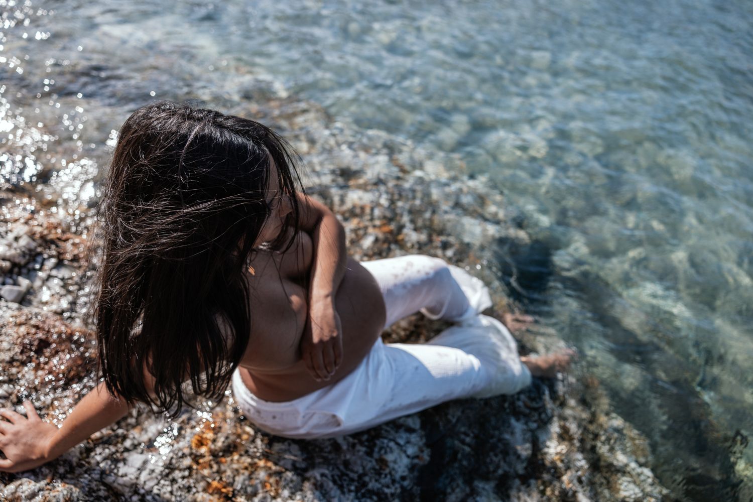 Pregnant woman in white pants sitting by crystal clear shallow ocean water.
