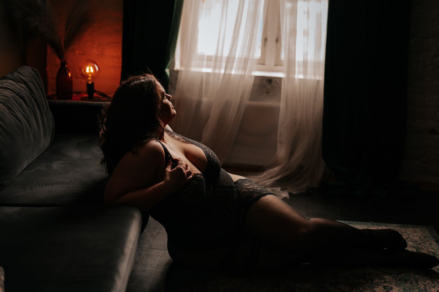 Woman in dark lace lingerie reclining on floor in warm moody light near a window and sofa.