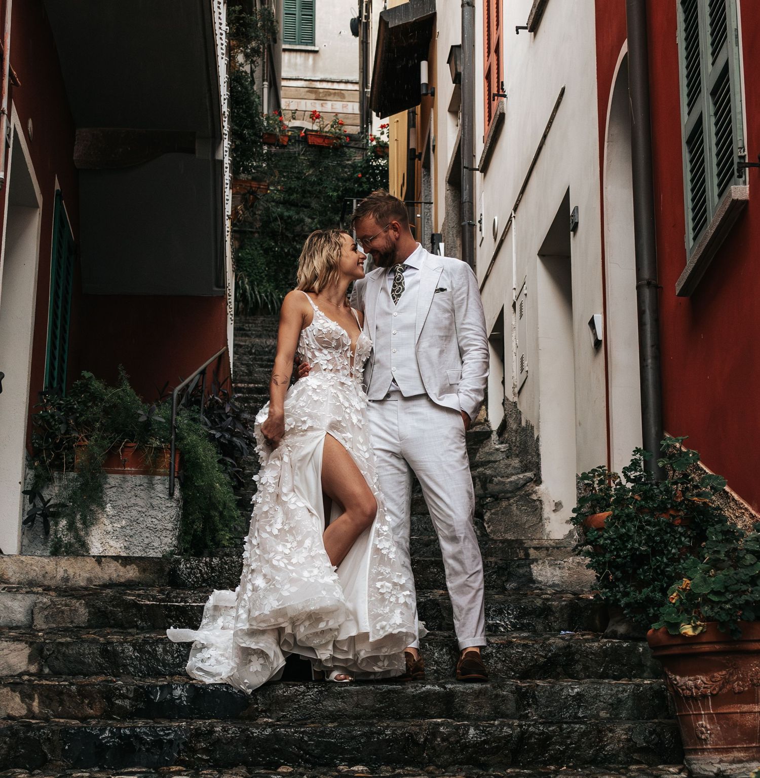 Elegant couple kissing on stone steps in Italian village, bride in floral gown, groom in white suit.