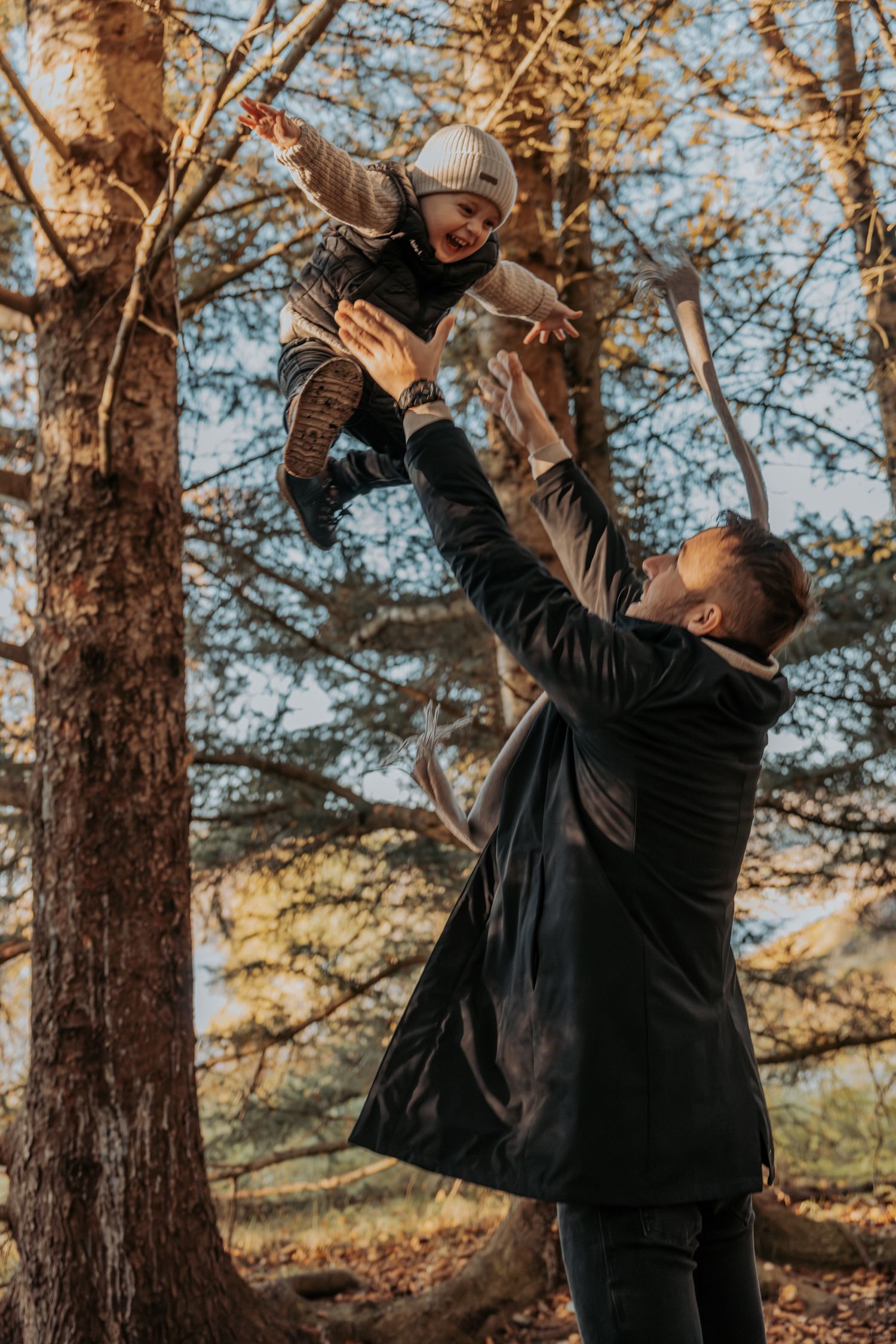 A father joyfully lifting his laughing toddler high in the air in a sunny autumn park.
