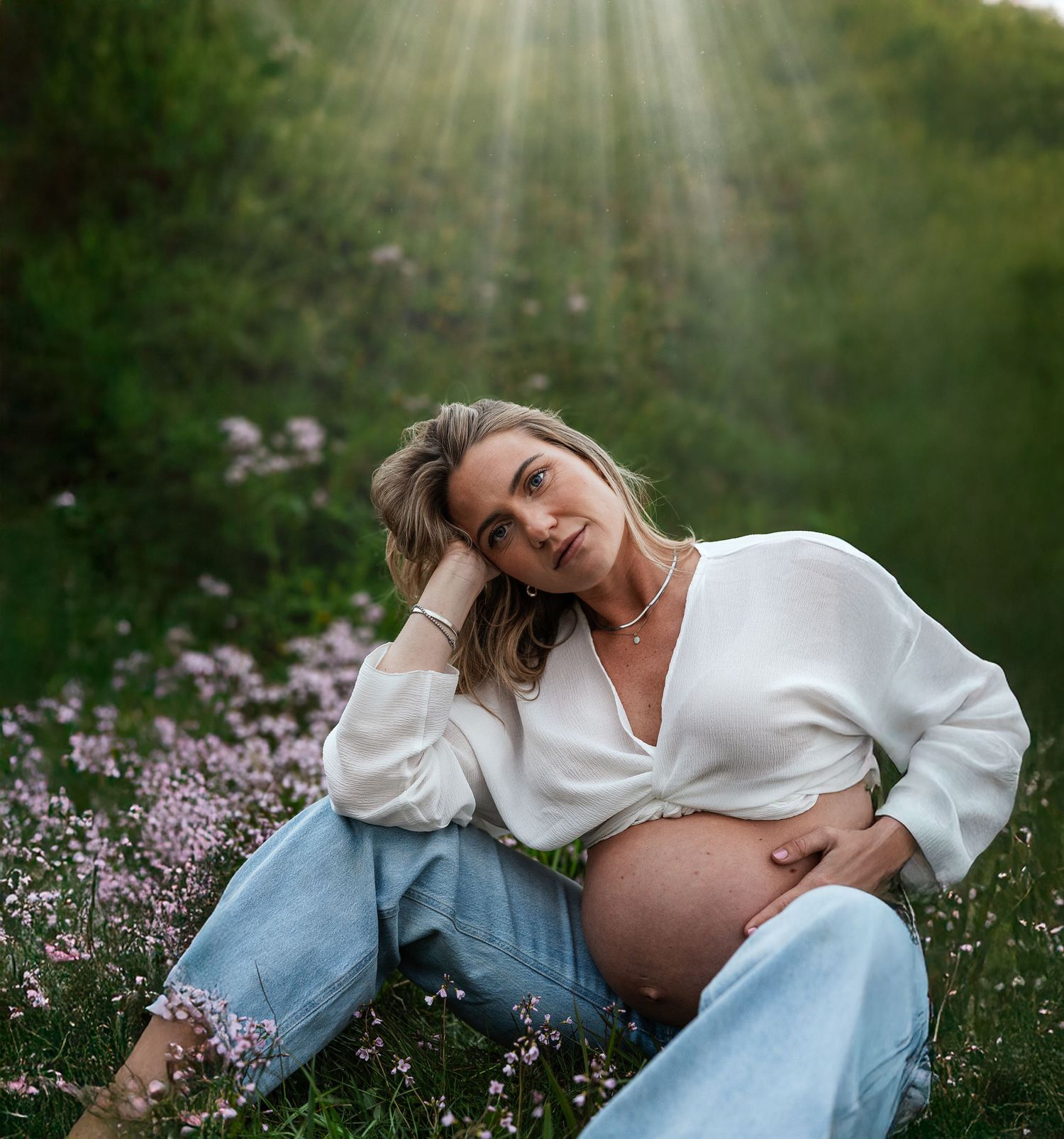 Pregnant woman in white blouse and jeans sitting in flower field with sunbeams, cradling baby bump outdoors.