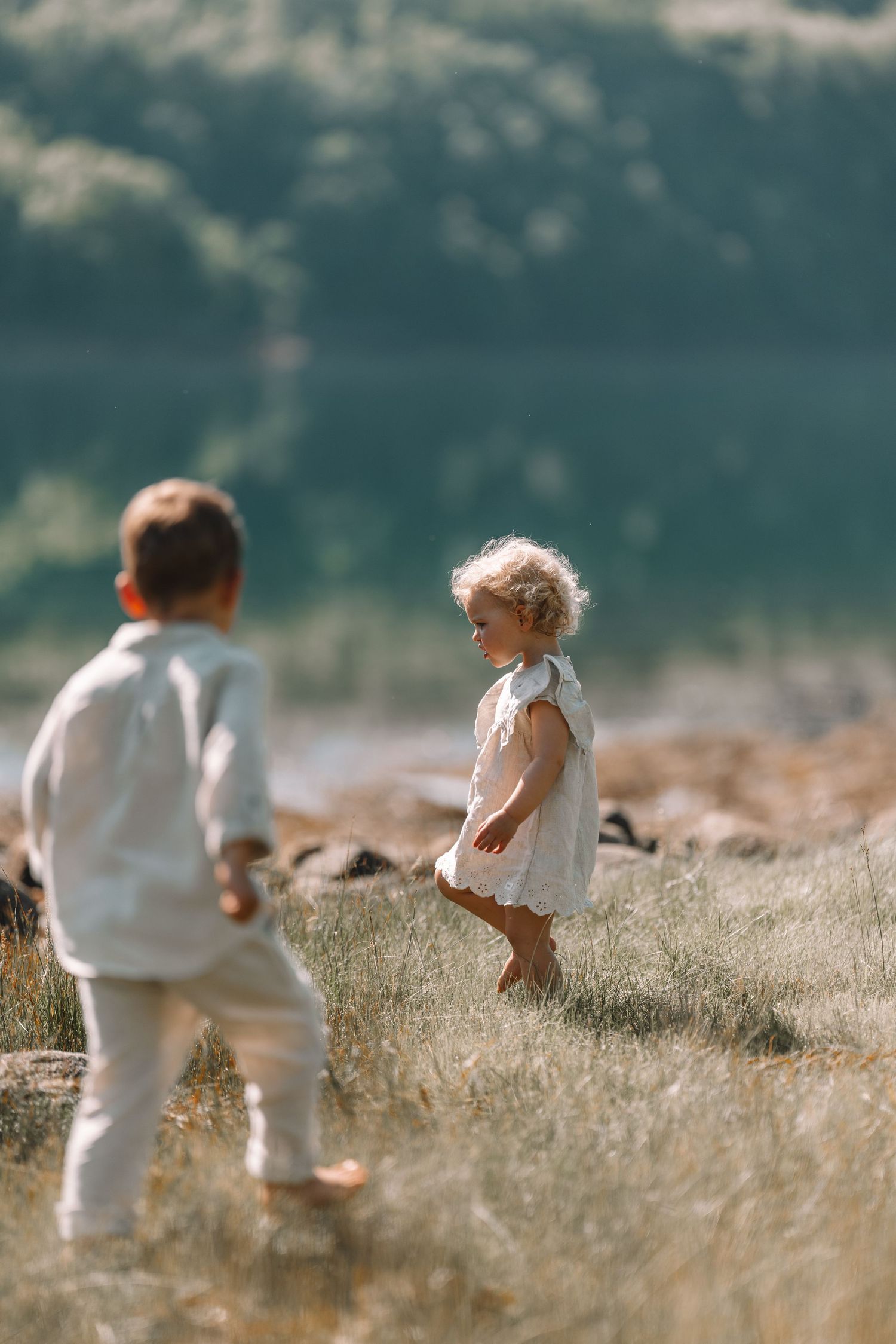 Two young children in white clothing play barefoot in a sunny meadow near a scenic lake.