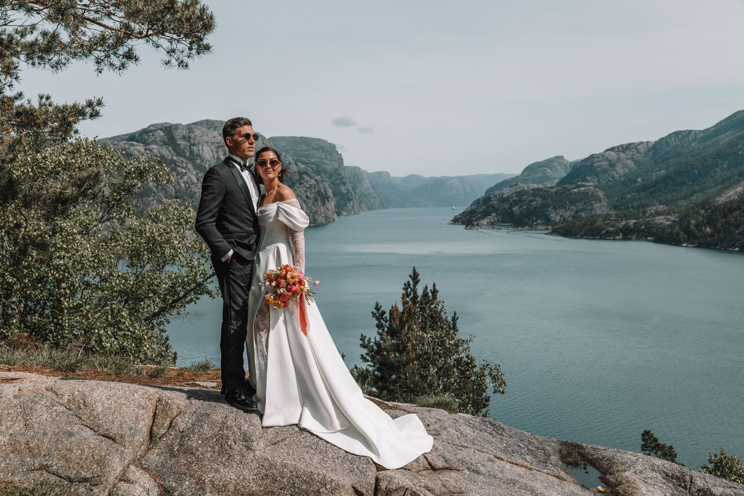 Bride and groom standing on rocky Norwegian fjord landscape with stunning turquoise water views behind them.