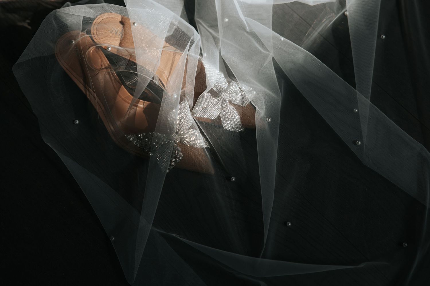 Artistic shot of bridal shoes and accessories draped under a flowing white veil with pearl details.
