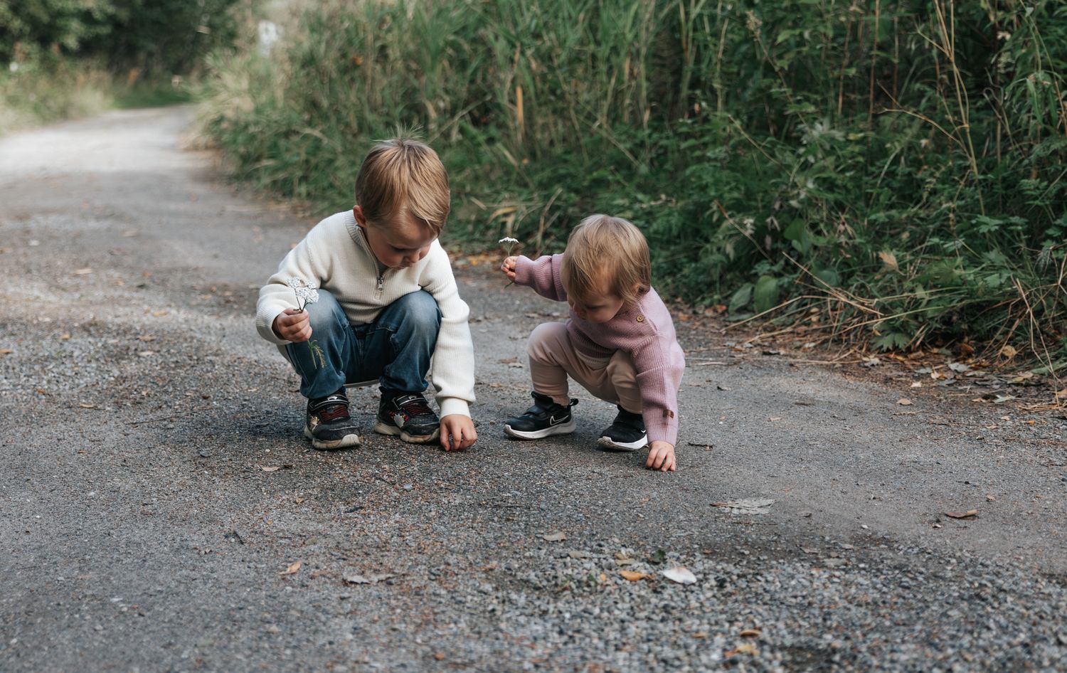 Two young children crouch on a gravel path exploring the ground, surrounded by lush green foliage.
