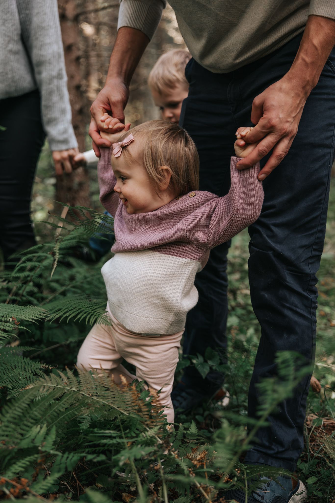 Baby girl in pink sweater learning to walk with family support among ferns.