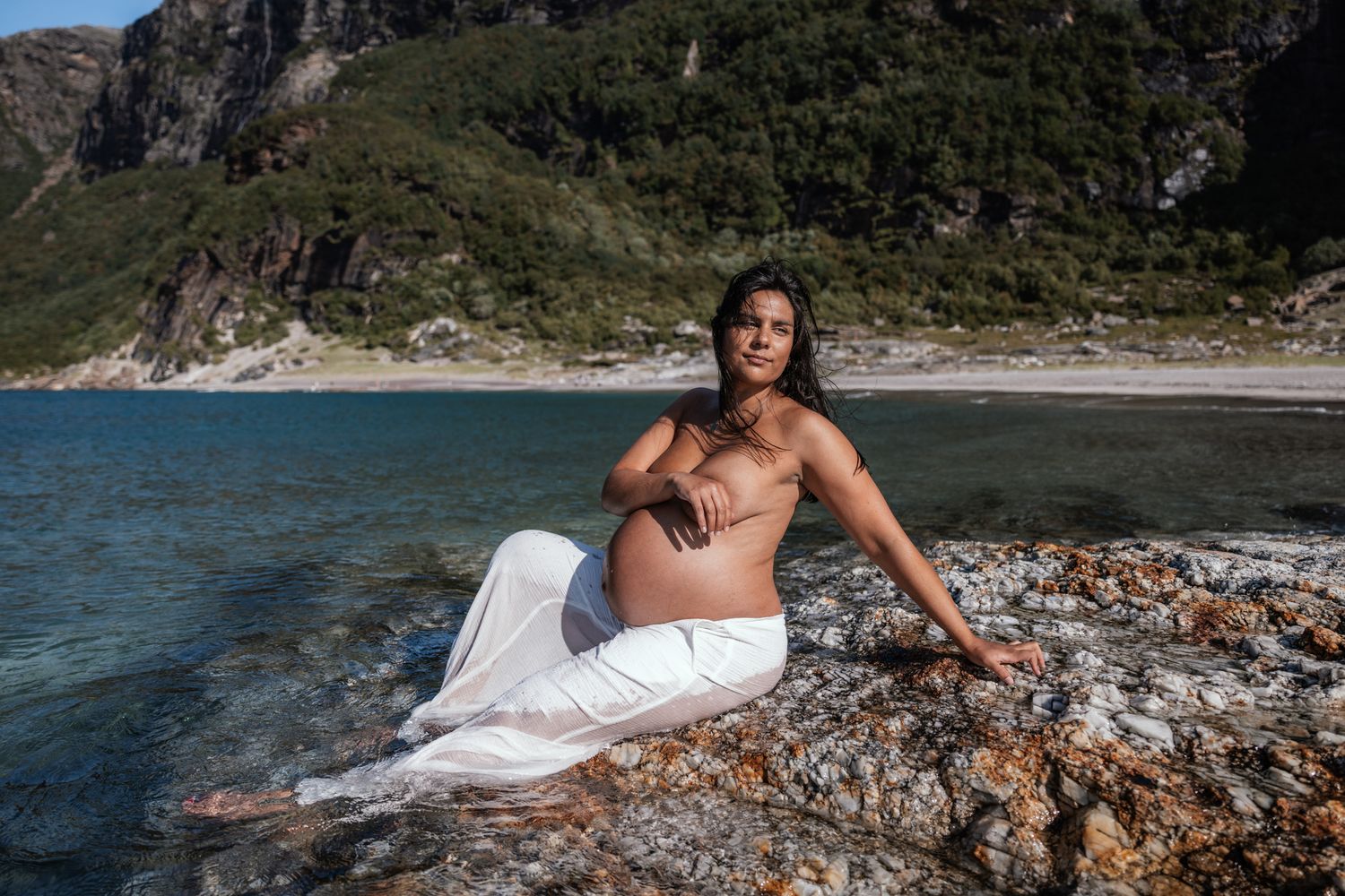 Pregnant woman in white pants sitting topless on rocks by scenic mountain lake.