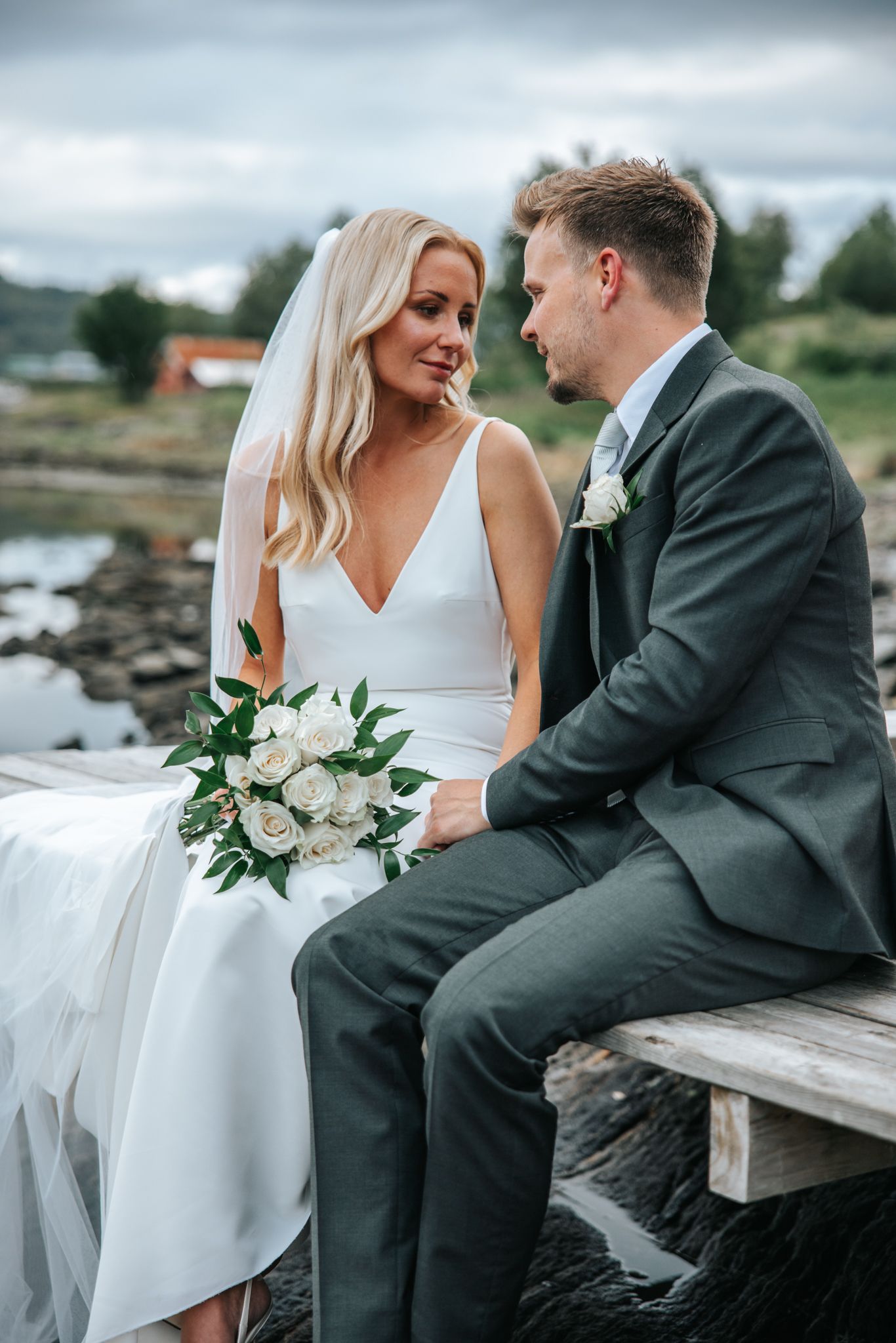 Bride and groom sitting on wooden dock by water, bride in sleek white gown holding white rose bouquet.
