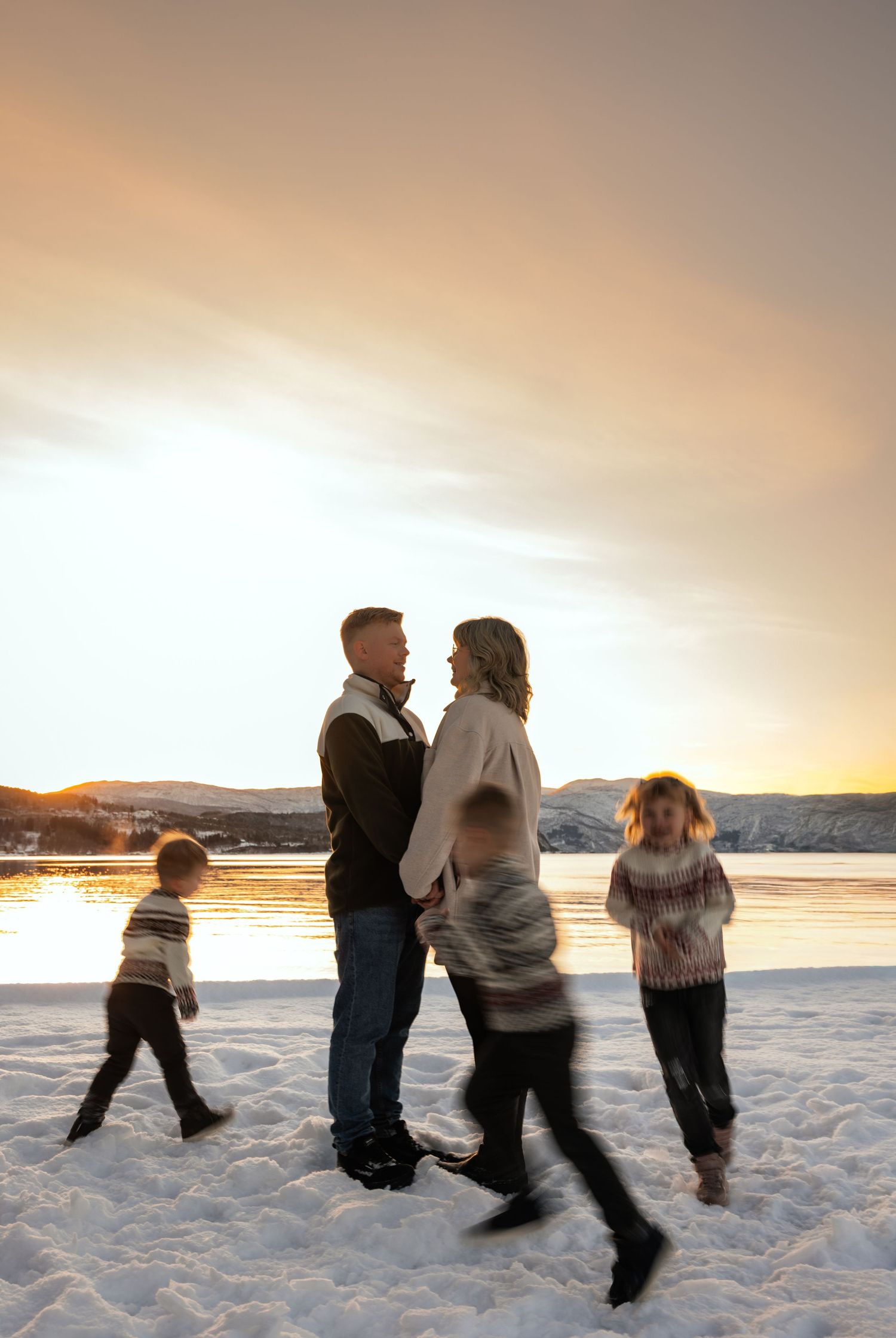 Family of four standing together on snowy landscape at sunset with mountains and water in background.