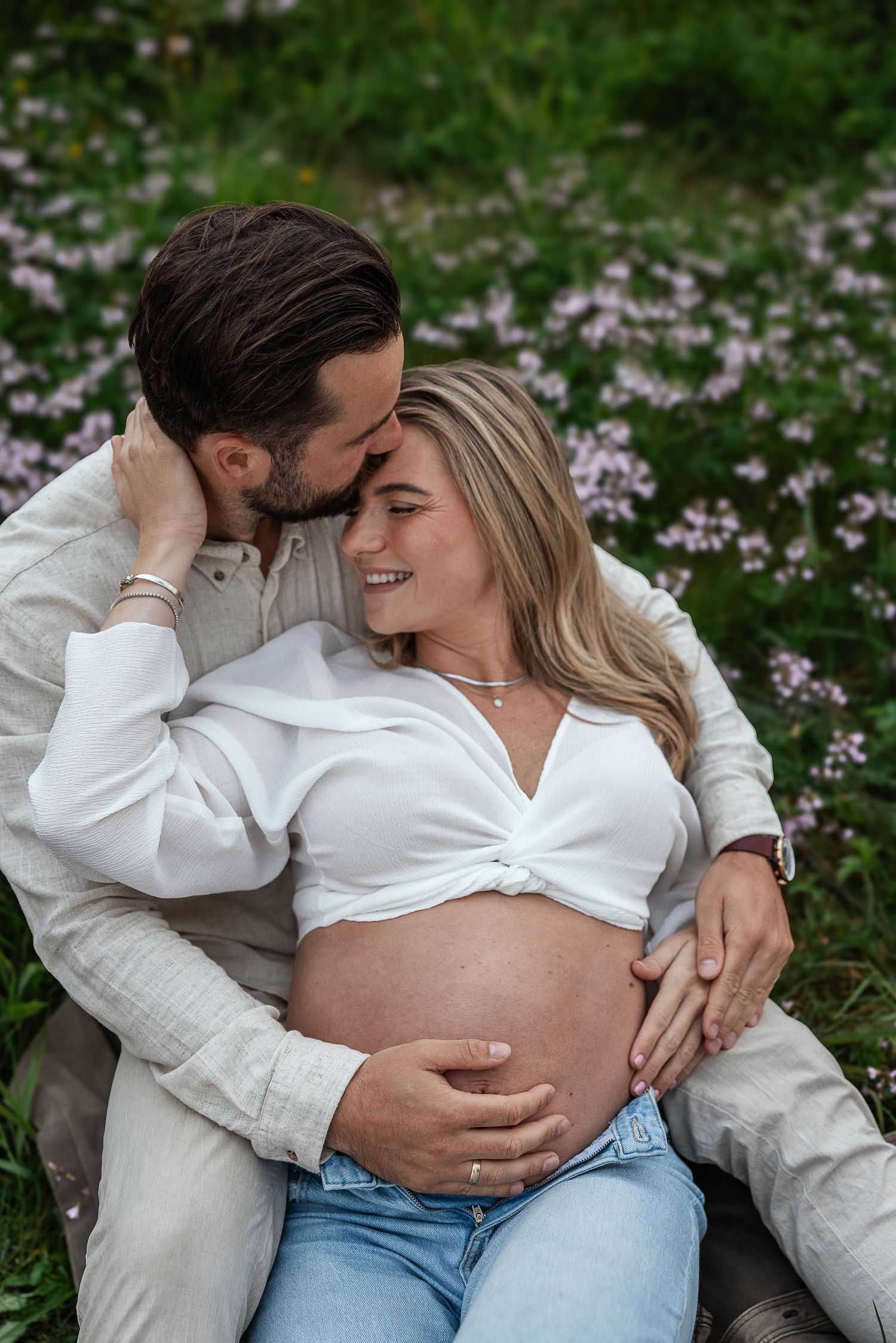 A happy couple sits in a flower field, man kissing pregnant woman's head as she smiles and cradles her baby bump.
