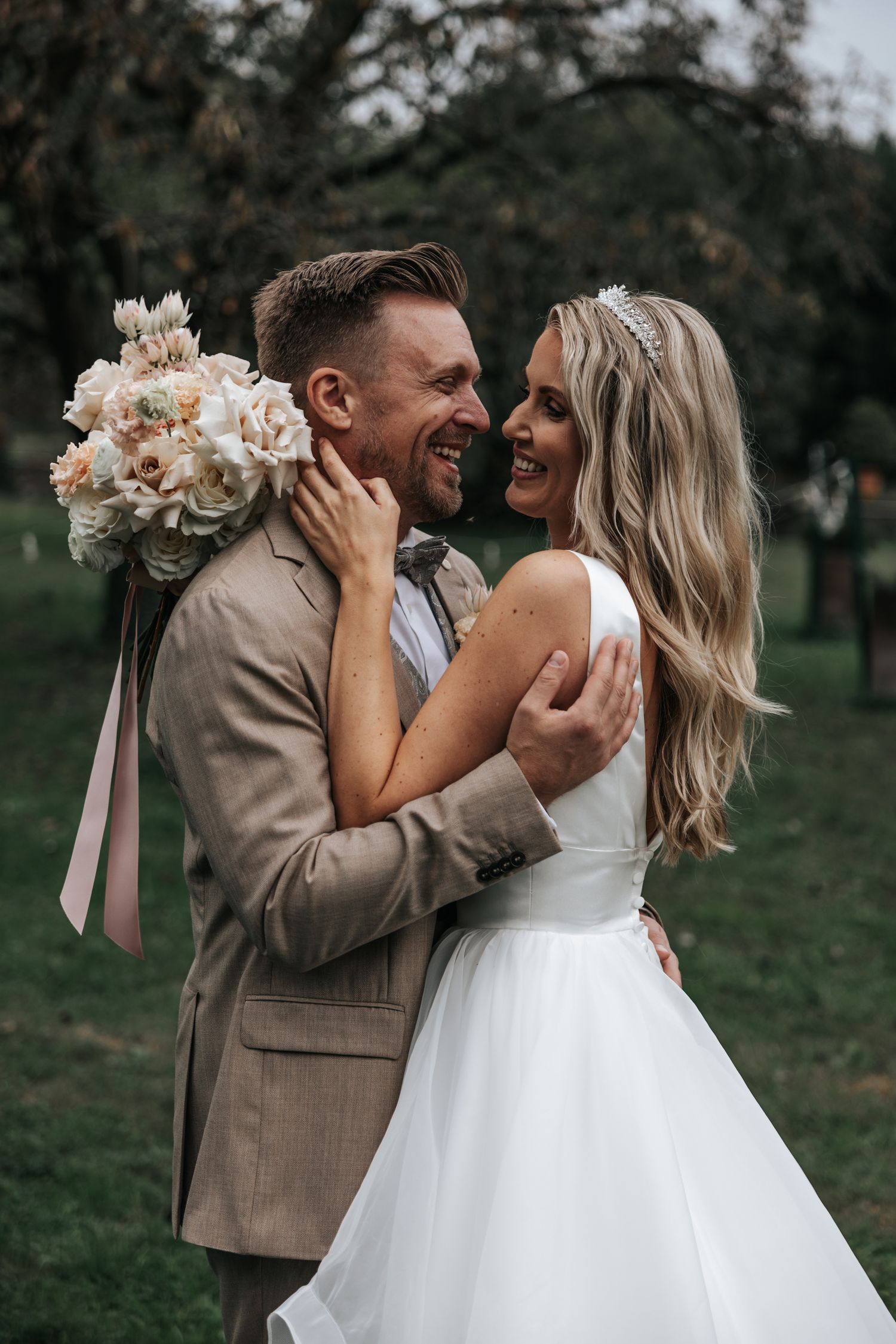 Joyful bride and groom embracing outdoors, bride holding floral bouquet, groom in tan suit, lush green background.