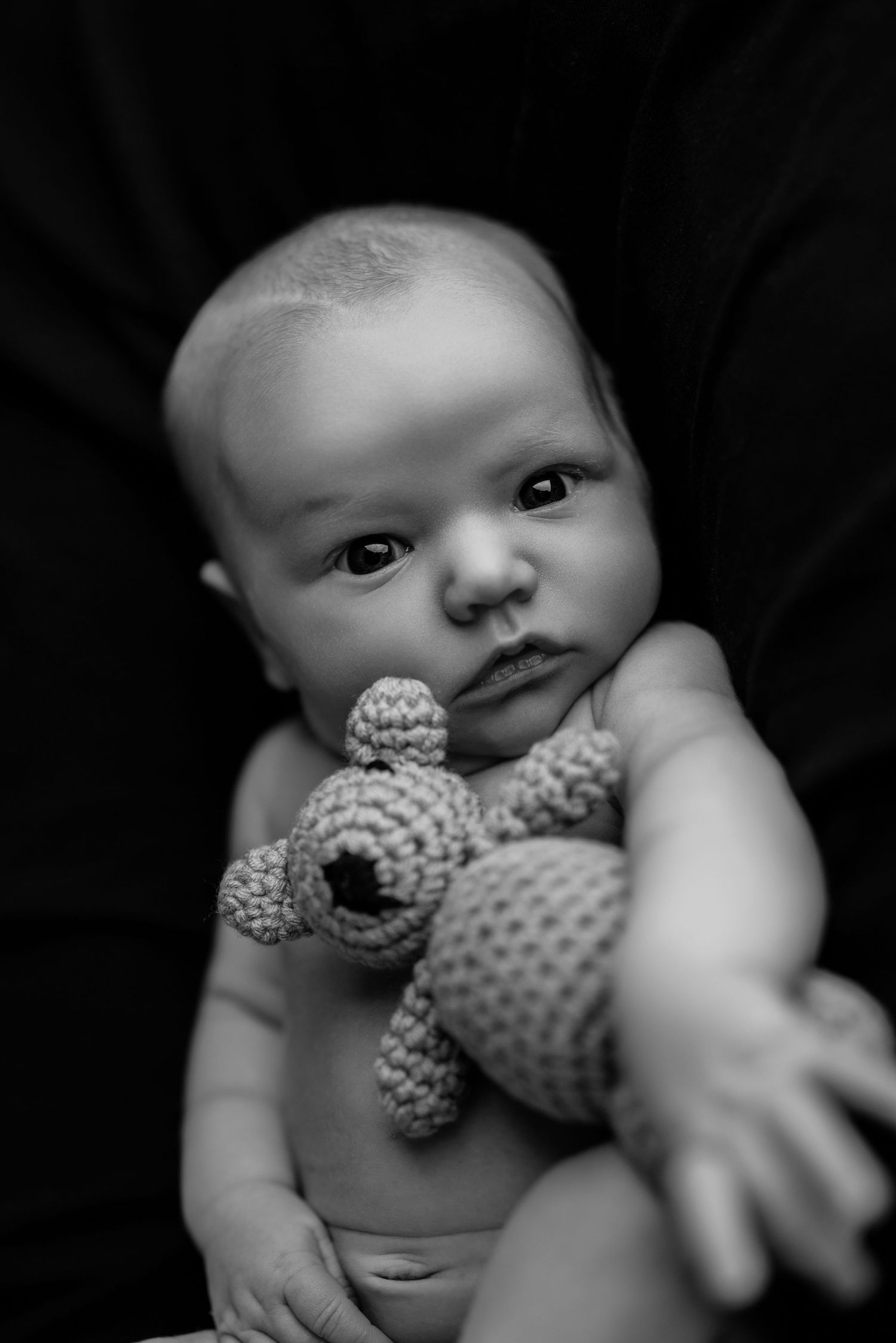 Black and white portrait of a baby holding a knitted teddy bear against a dark background.